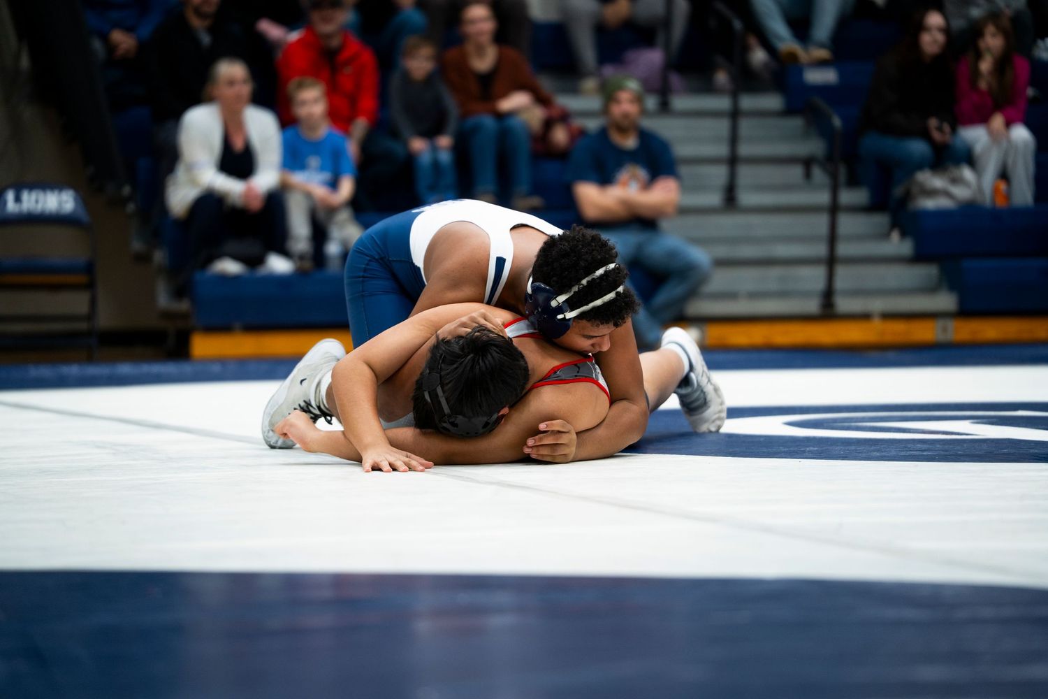 High school wrestler in blue pins opponent face-down on wrestling mat during competitive match.
