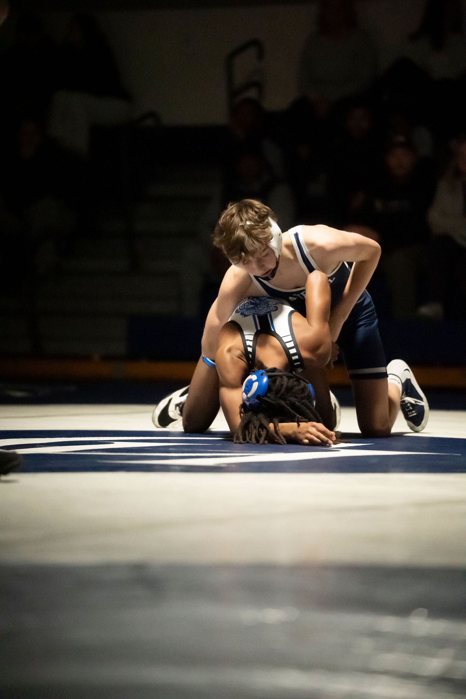 High school wrestler in white singlet dominates opponent on the mat during an indoor wrestling competition.