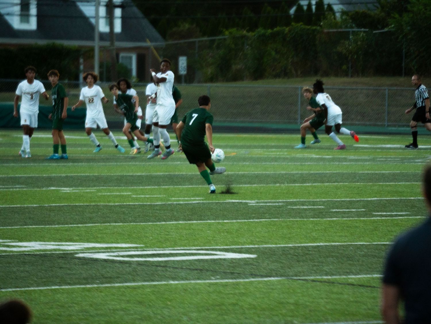 Trinity #7 advances soccer ball downfield while teammates and opponents compete on green turf field.