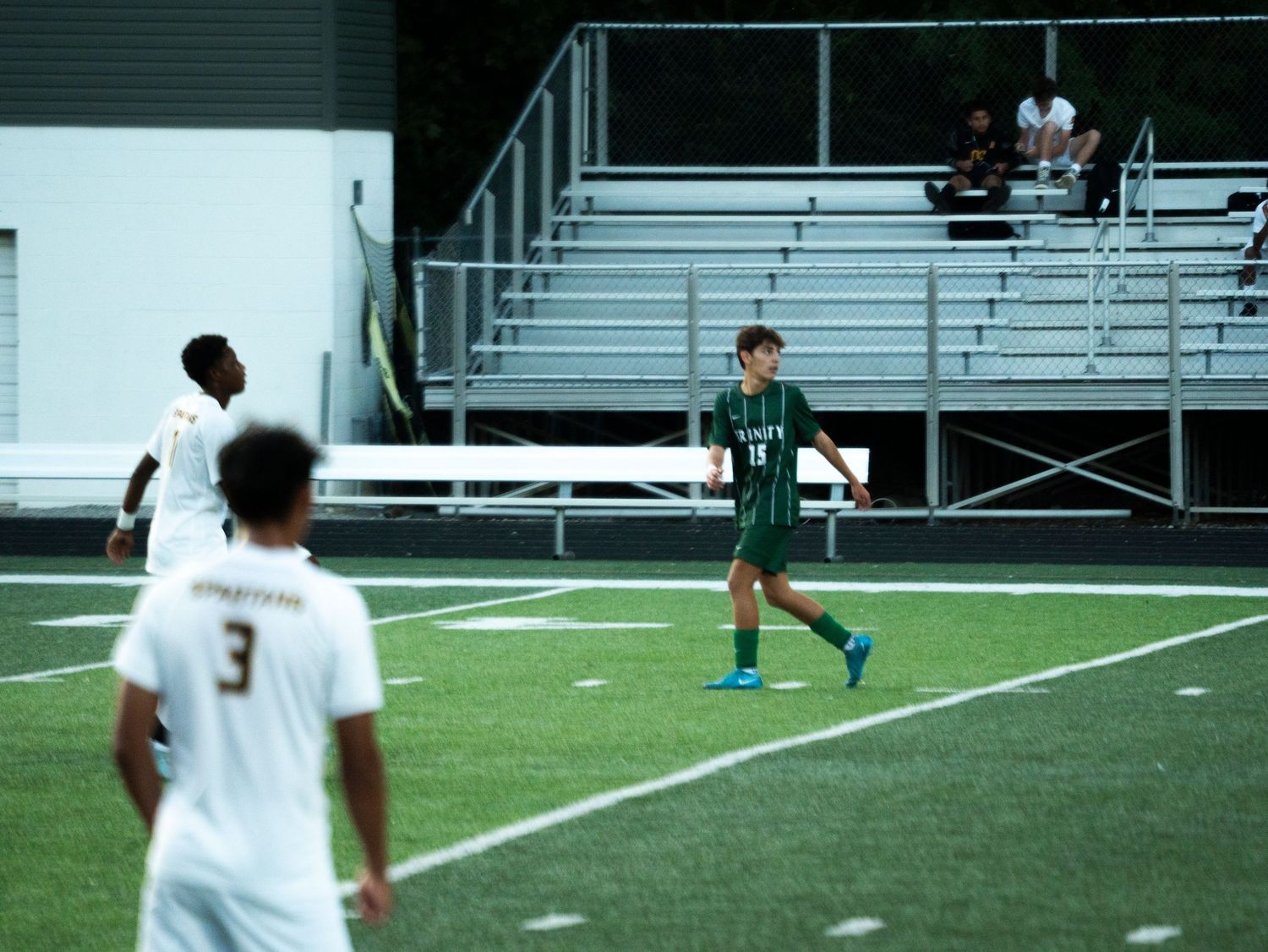 Trinity soccer player #11 walks on turf field during evening match with opponent #3 in foreground.