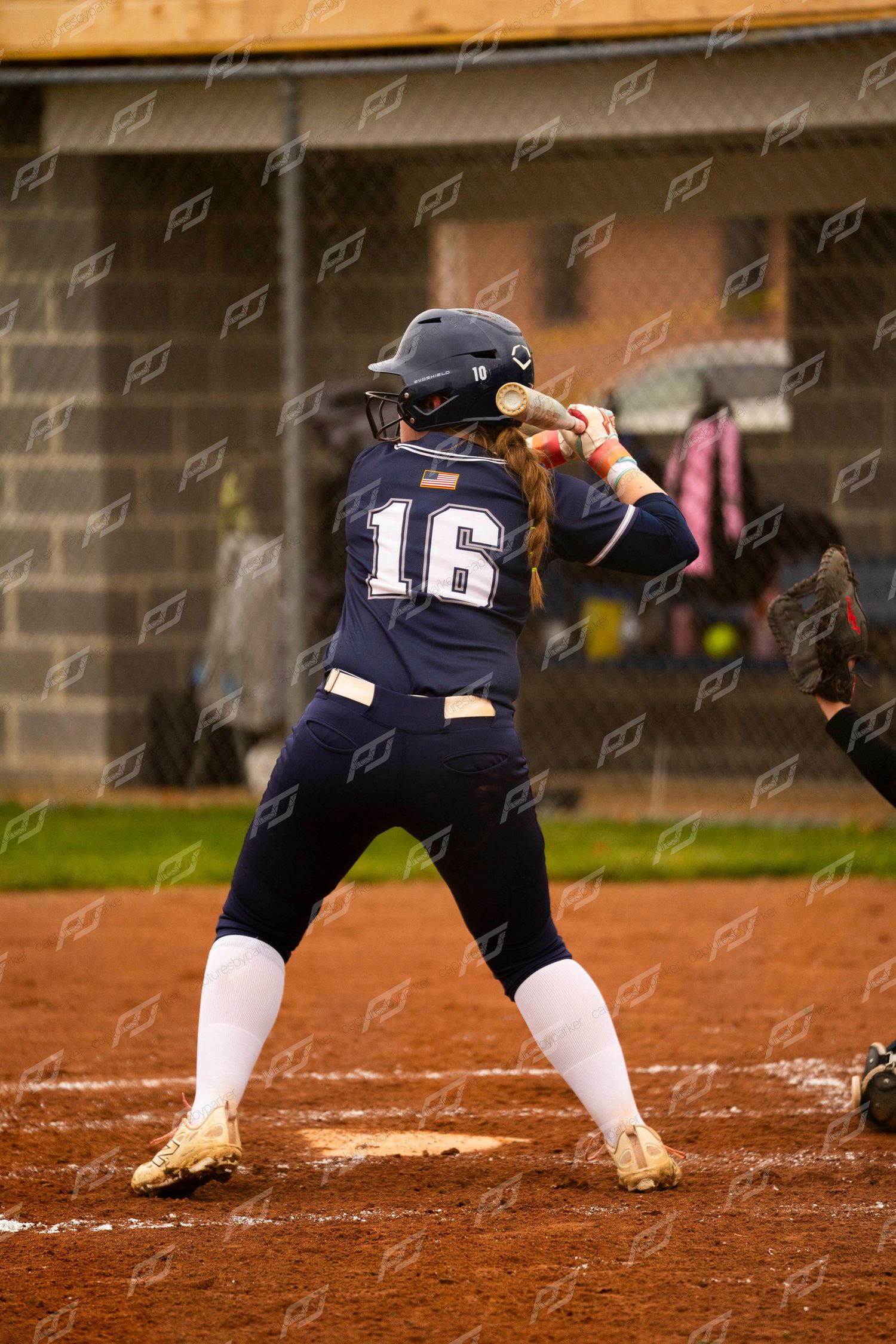 Softball player #16 in navy uniform stands at bat in ready stance on dirt infield during game.