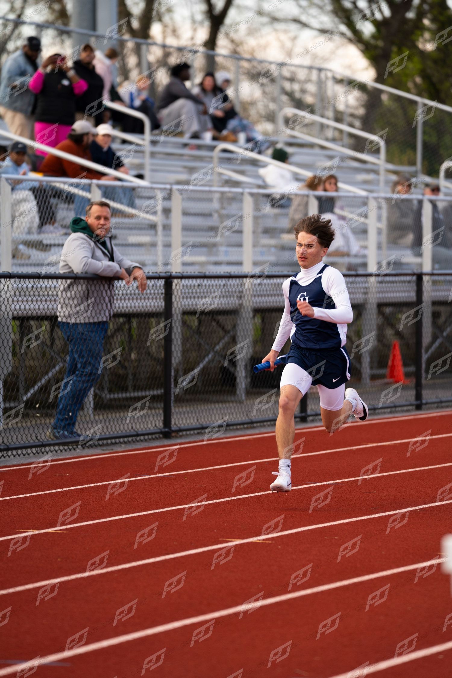 Young male athlete in navy uniform runs with blue baton on red track during high school relay race.