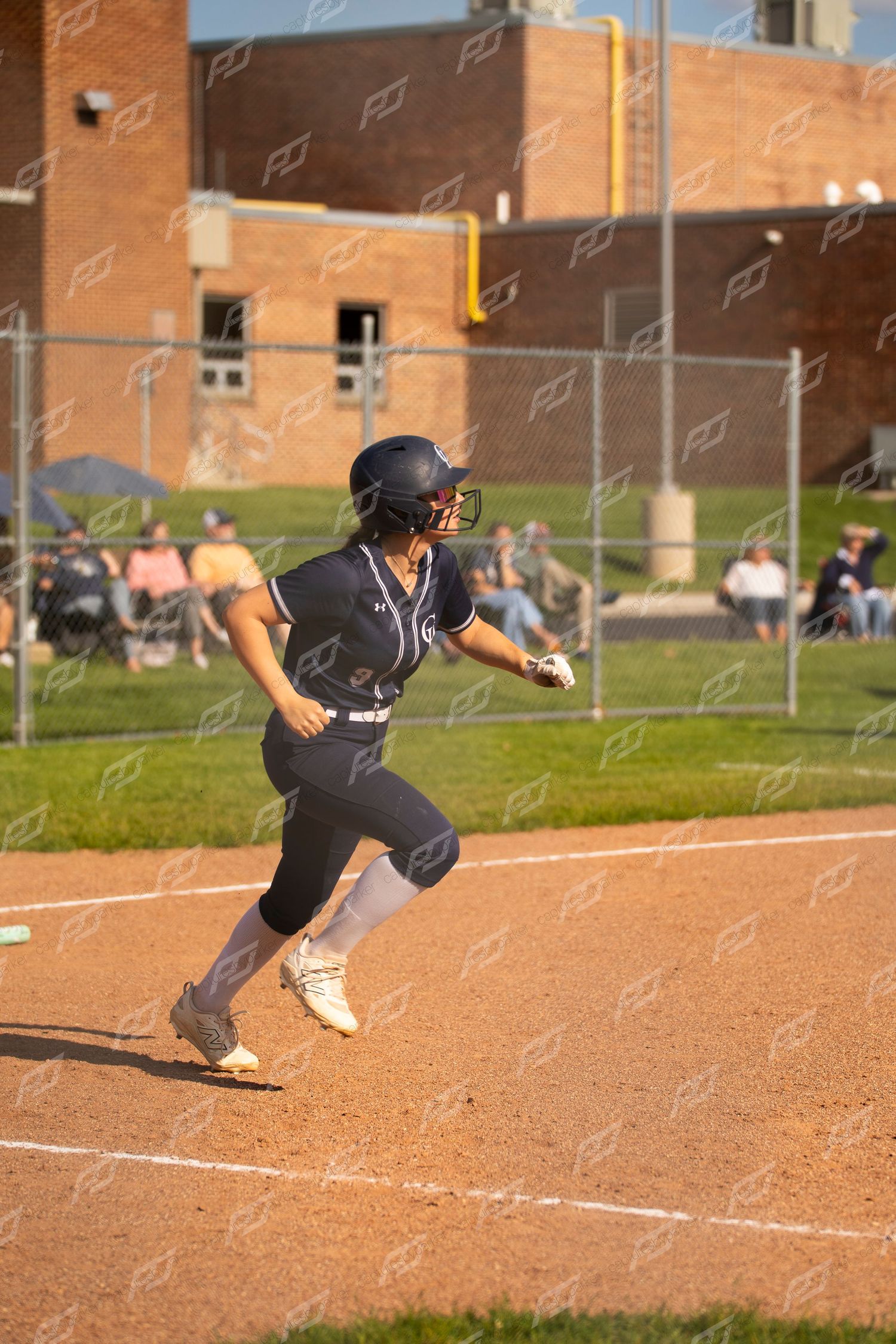 Female softball player in navy uniform sprints toward home plate during evening game.