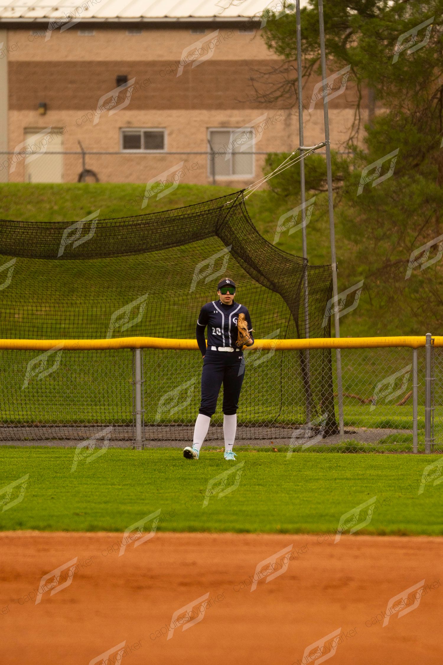 Softball outfielder in navy uniform standing near the warning track fence during an outdoor game.