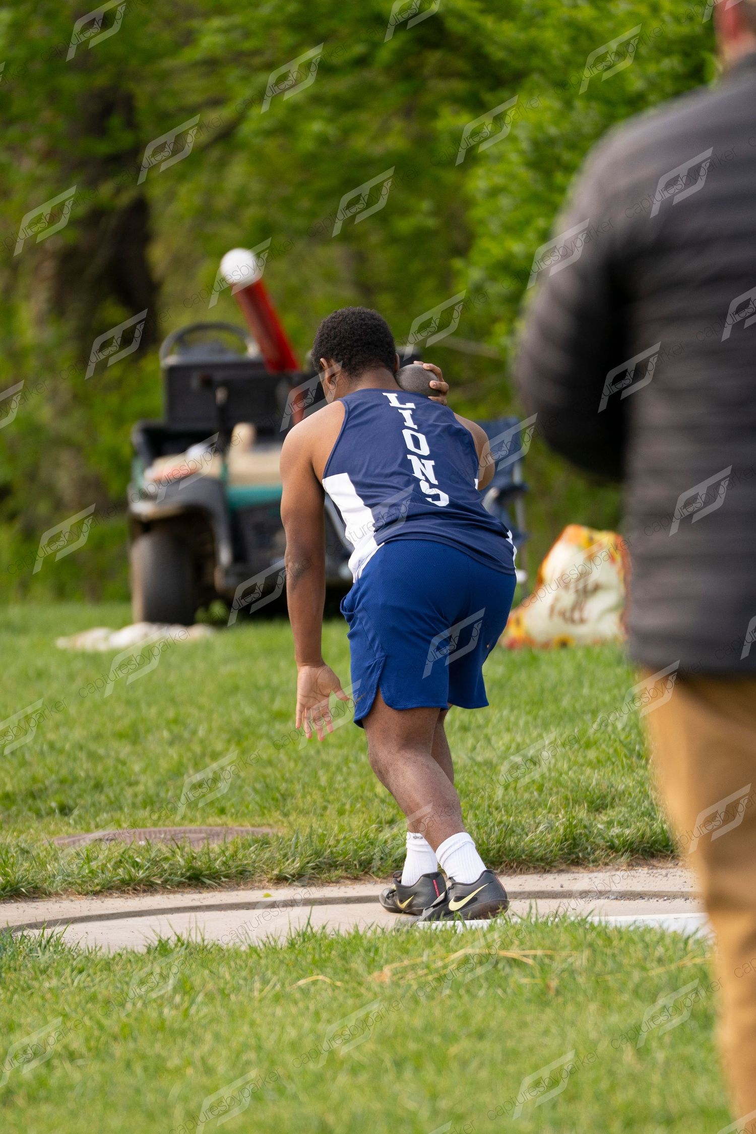 Male athlete in blue LIONS uniform releasing shot put during outdoor track and field competition.