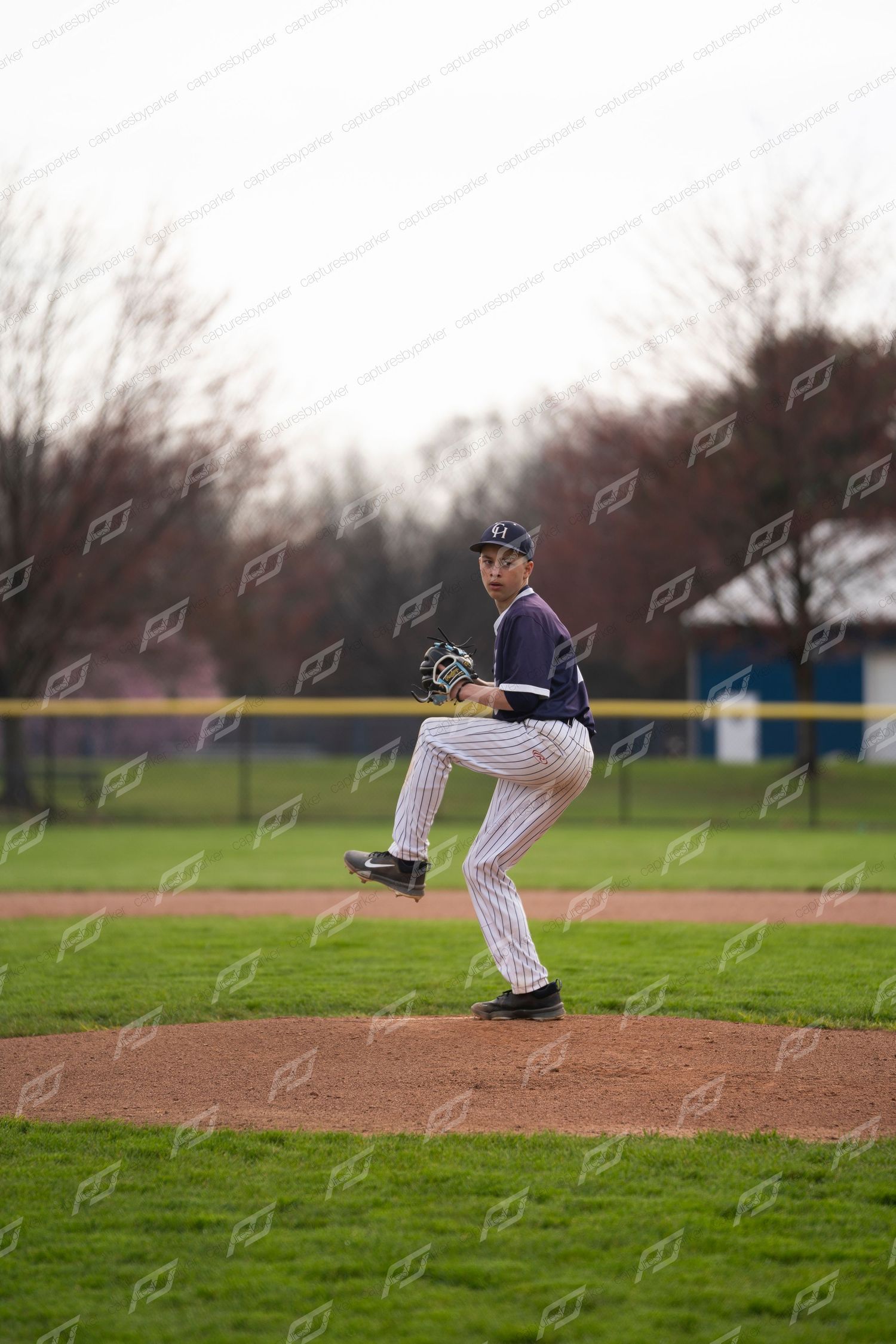 Youth baseball pitcher in navy pinstripe uniform in wind-up position on the mound during a spring game.