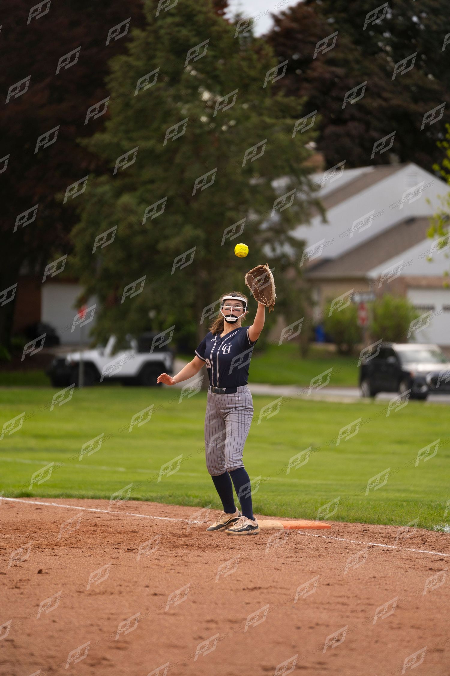 Softball player catches a yellow ball in her glove during an outdoor game on a dirt infield.