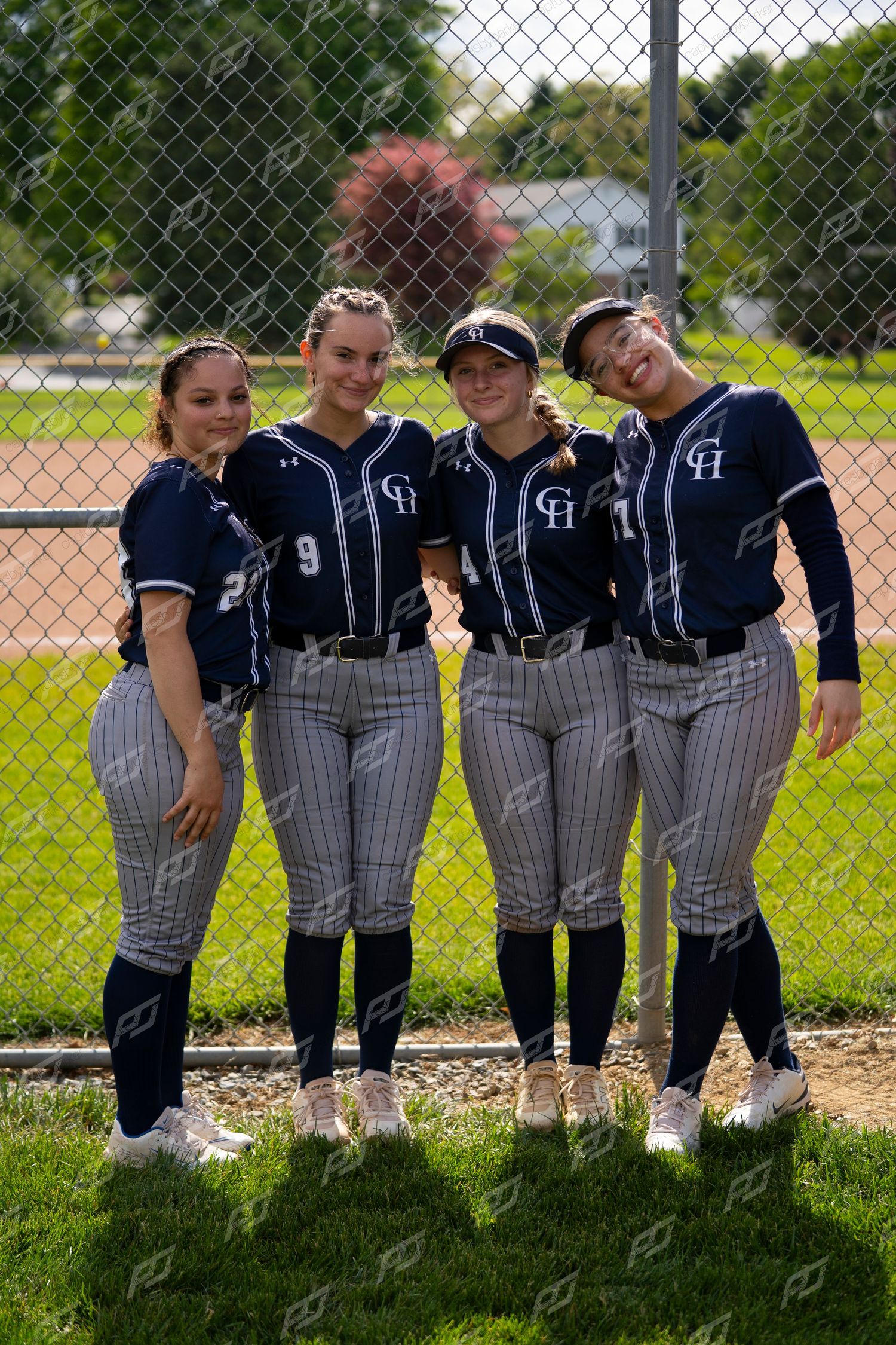 Four female softball players in navy pinstripe uniforms pose together smiling in front of a chain-link fence.