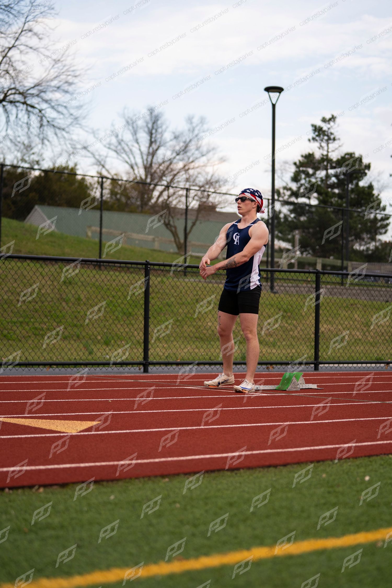 Male track athlete in navy uniform stands at starting blocks on red track looking ahead.