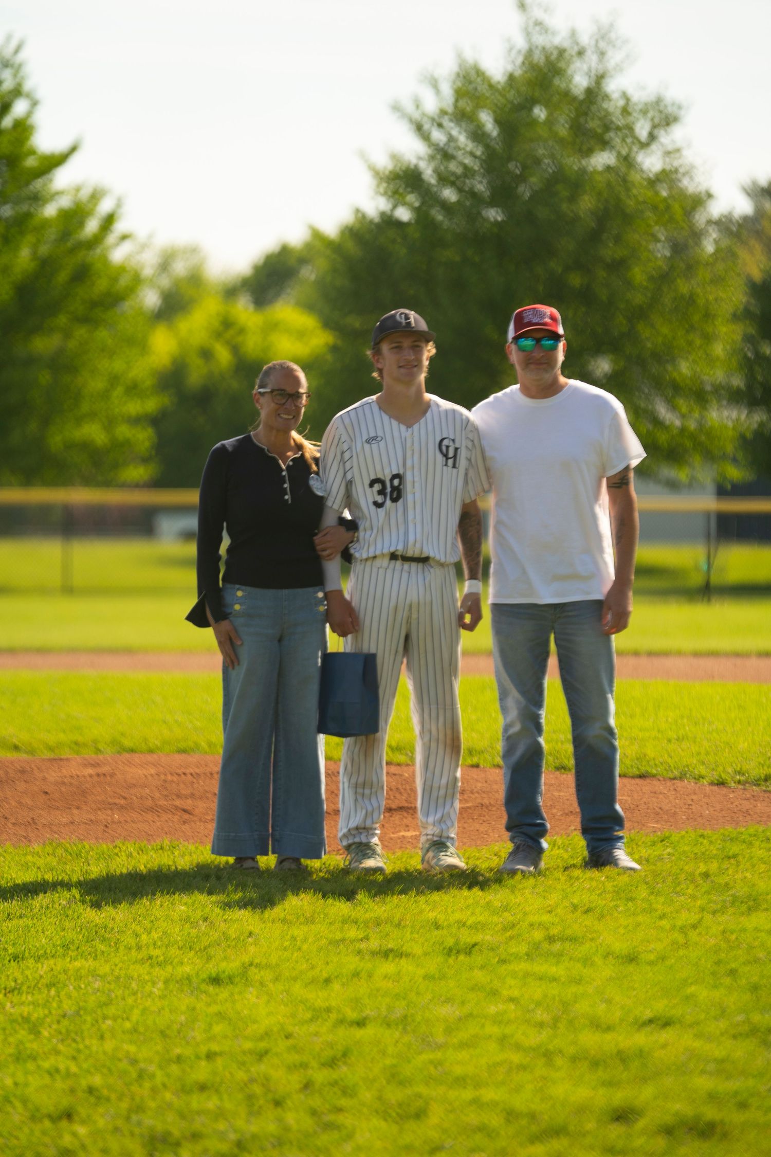 Baseball player #38 in pinstripe uniform poses with two parents holding a gift on a sunny baseball field.