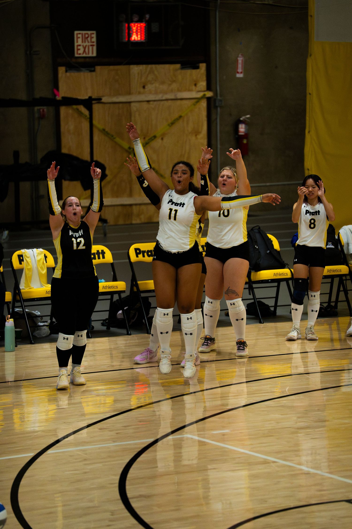 Pratt volleyball players celebrate with arms raised during a match on a hardwood court, wearing black and white uniforms.