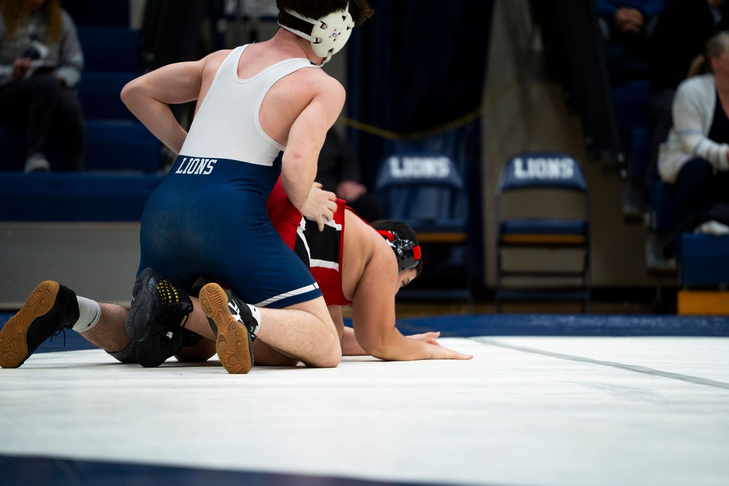 A wrestler in blue pins opponent on white mat during high school wrestling match in Lions gymnasium.