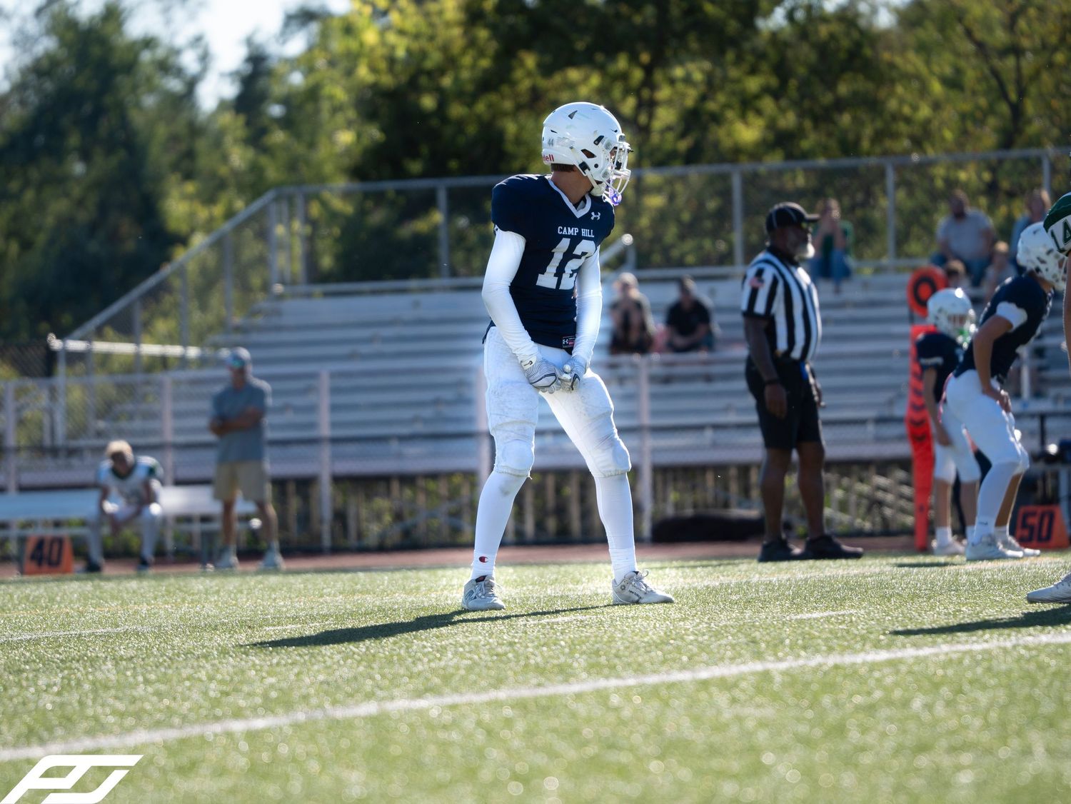 Youth football player in navy uniform stands in ready position on turf field with referee nearby.