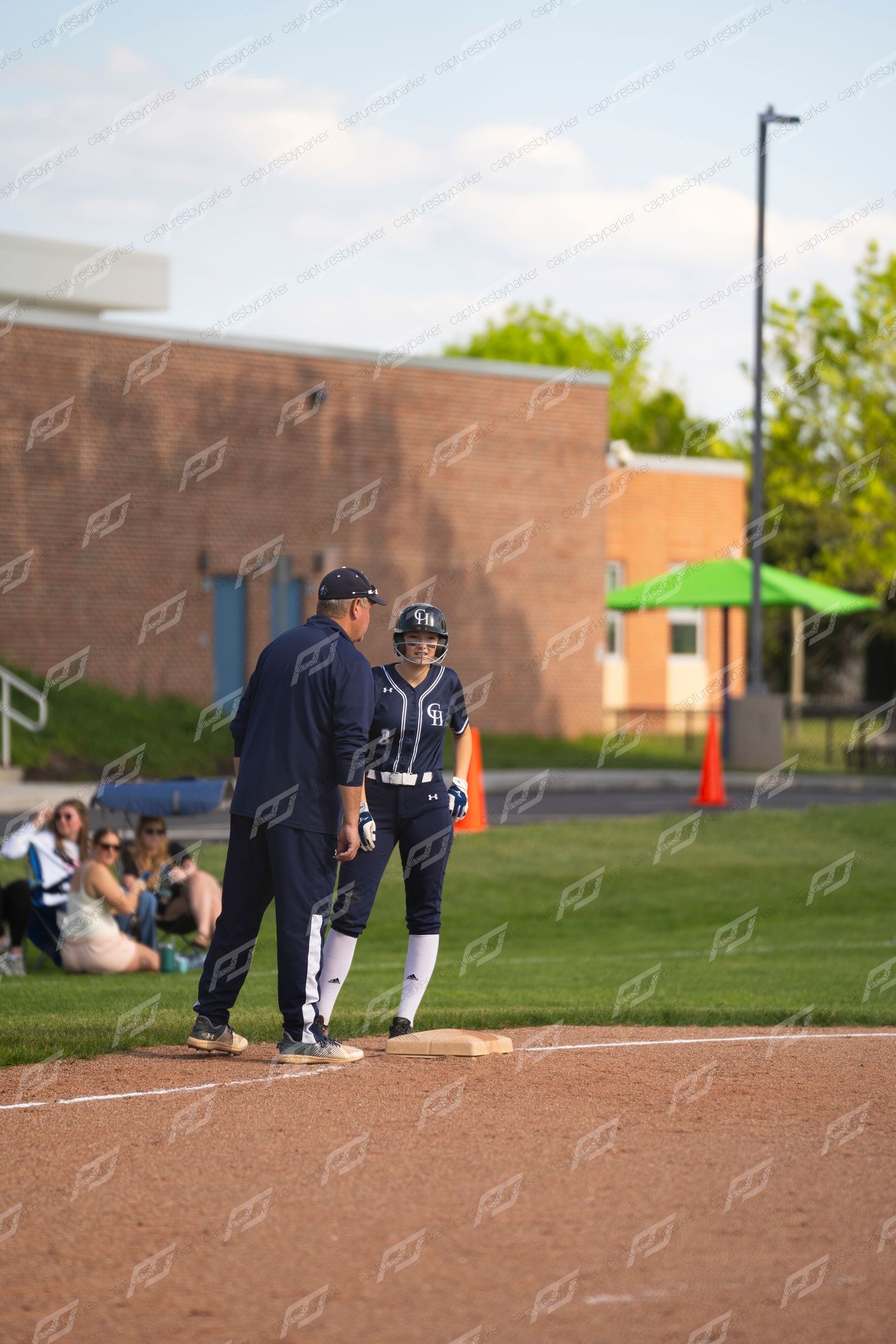 A softball coach in navy uniform talks with a player at first base during an evening game.