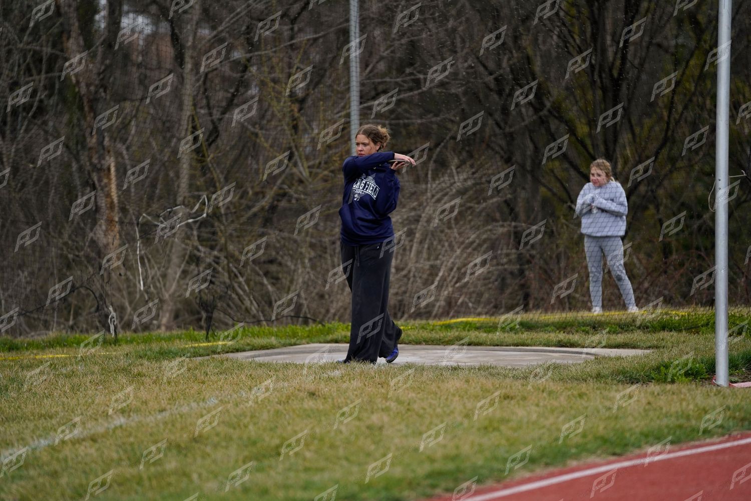 Female athlete in navy jacket releases a discus during an outdoor high school track and field meet.