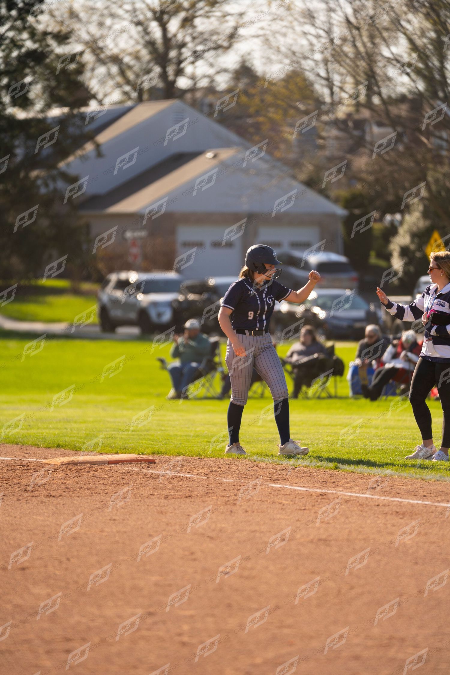 Female softball player in navy pinstripe uniform celebrating at third base during outdoor game.