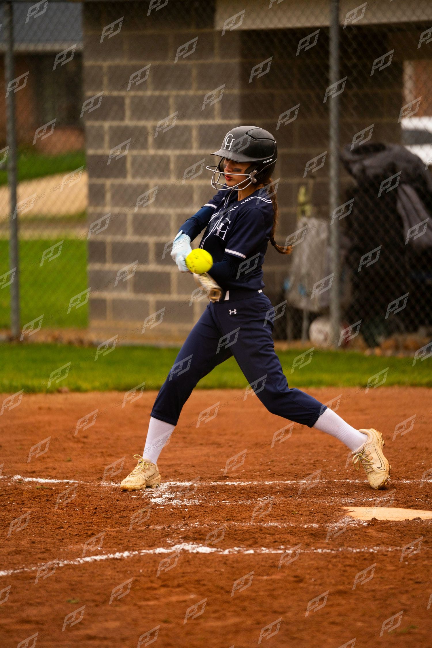 Female softball player in navy uniform makes contact with yellow ball during competitive game.