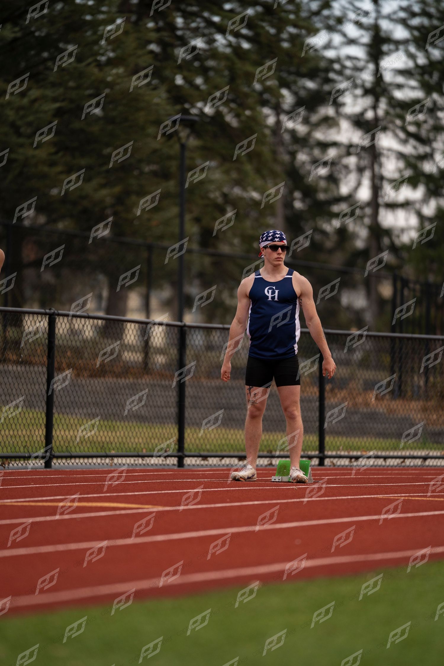 Male track athlete in navy uniform stands on red track wearing sunglasses.