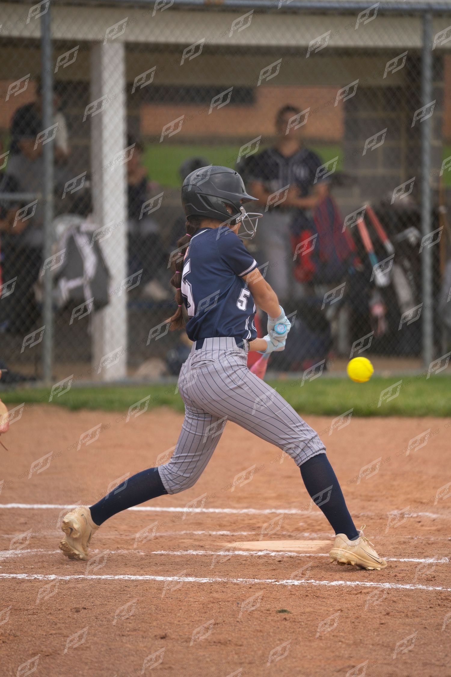 Female softball batter in navy pinstripe uniform swings at a yellow pitch during a game.