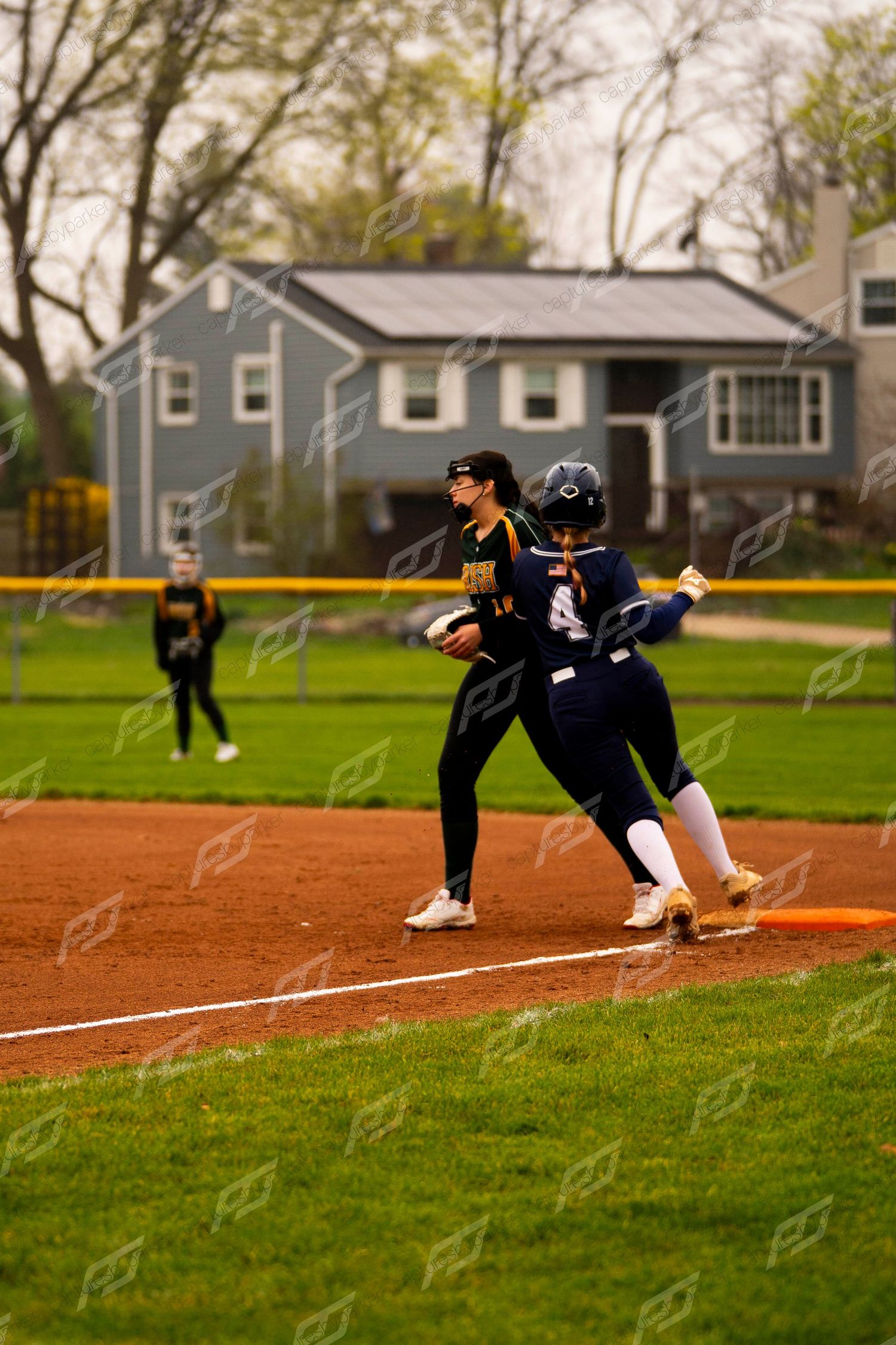 Two softball players collide at first base during a game on a clay infield with a blue house in the background.