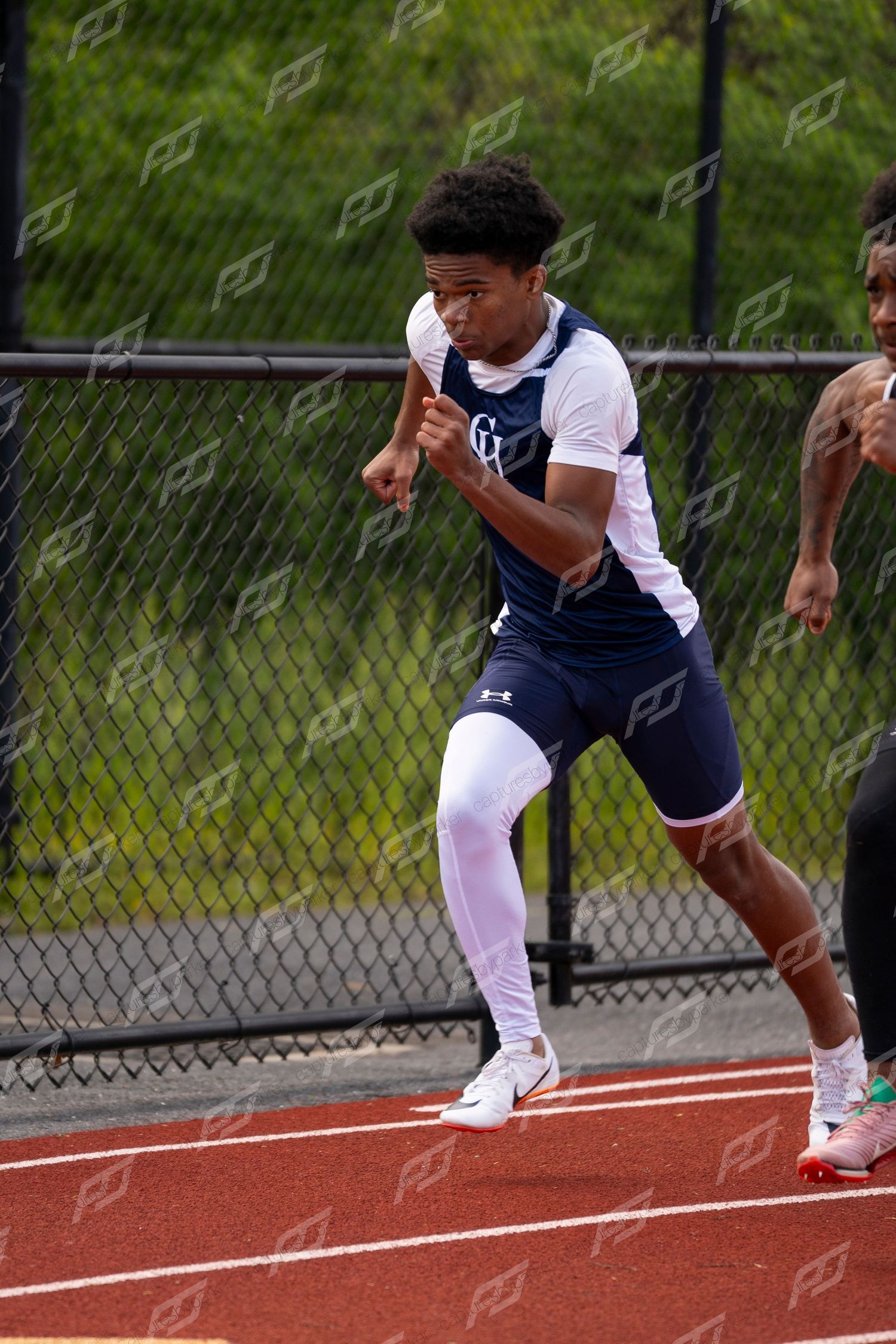Young male sprinter in navy uniform accelerates on red track during outdoor track and field meet.