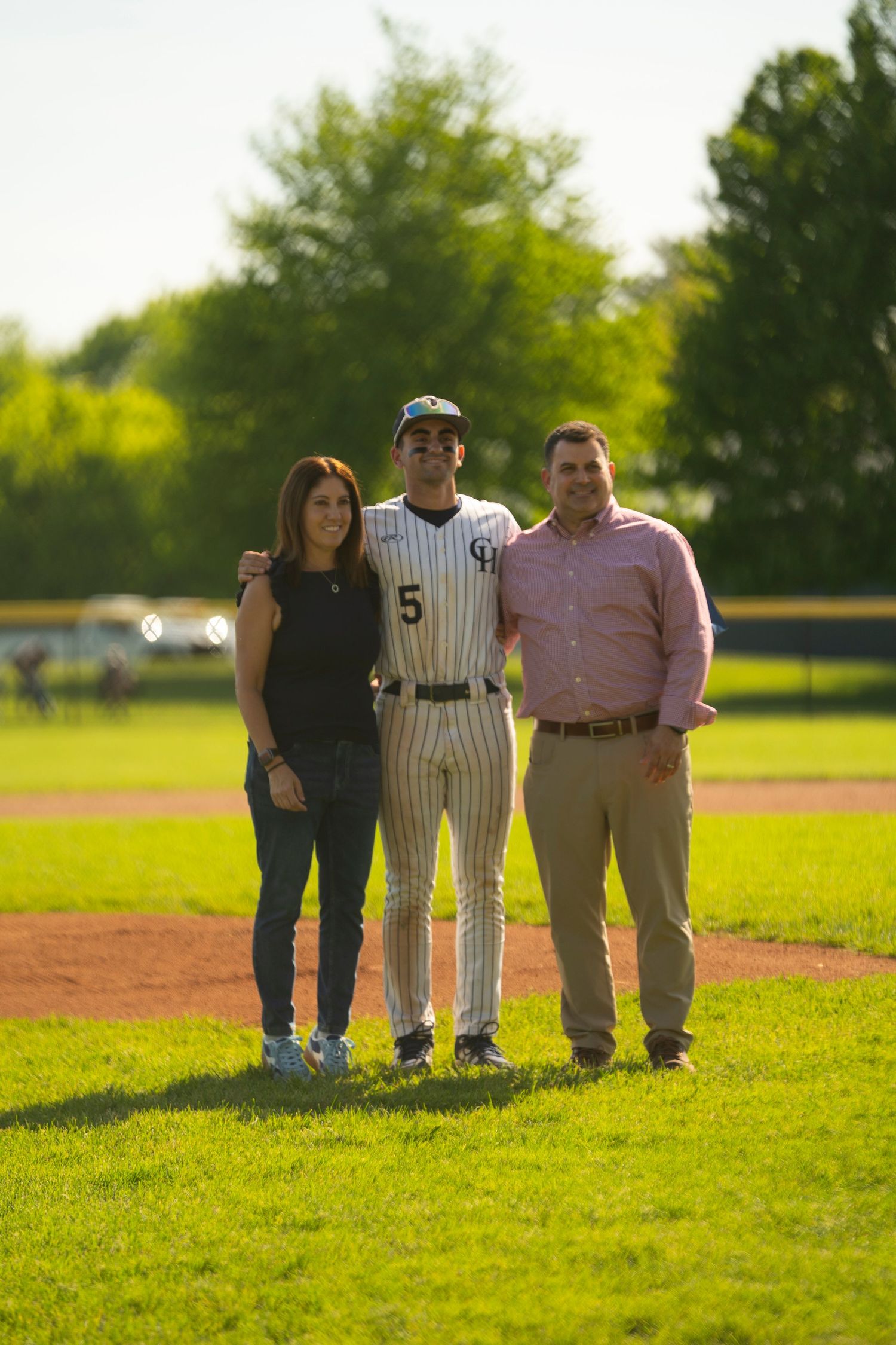 Baseball player #5 in pinstripe uniform poses with two parents on a bright sunny green baseball field.