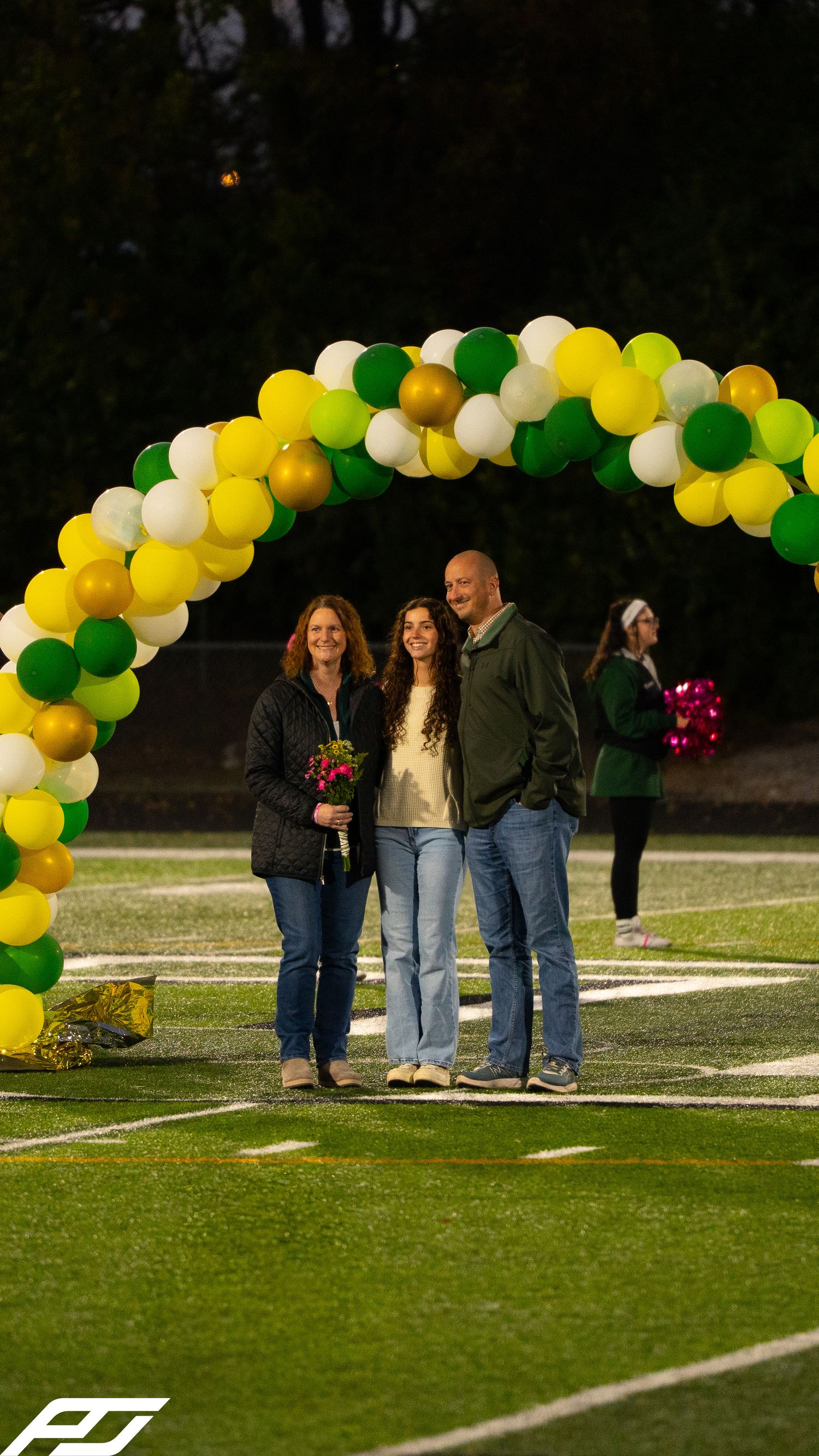 Family of three smiles under a festive green and yellow balloon arch during a school sports senior night event.