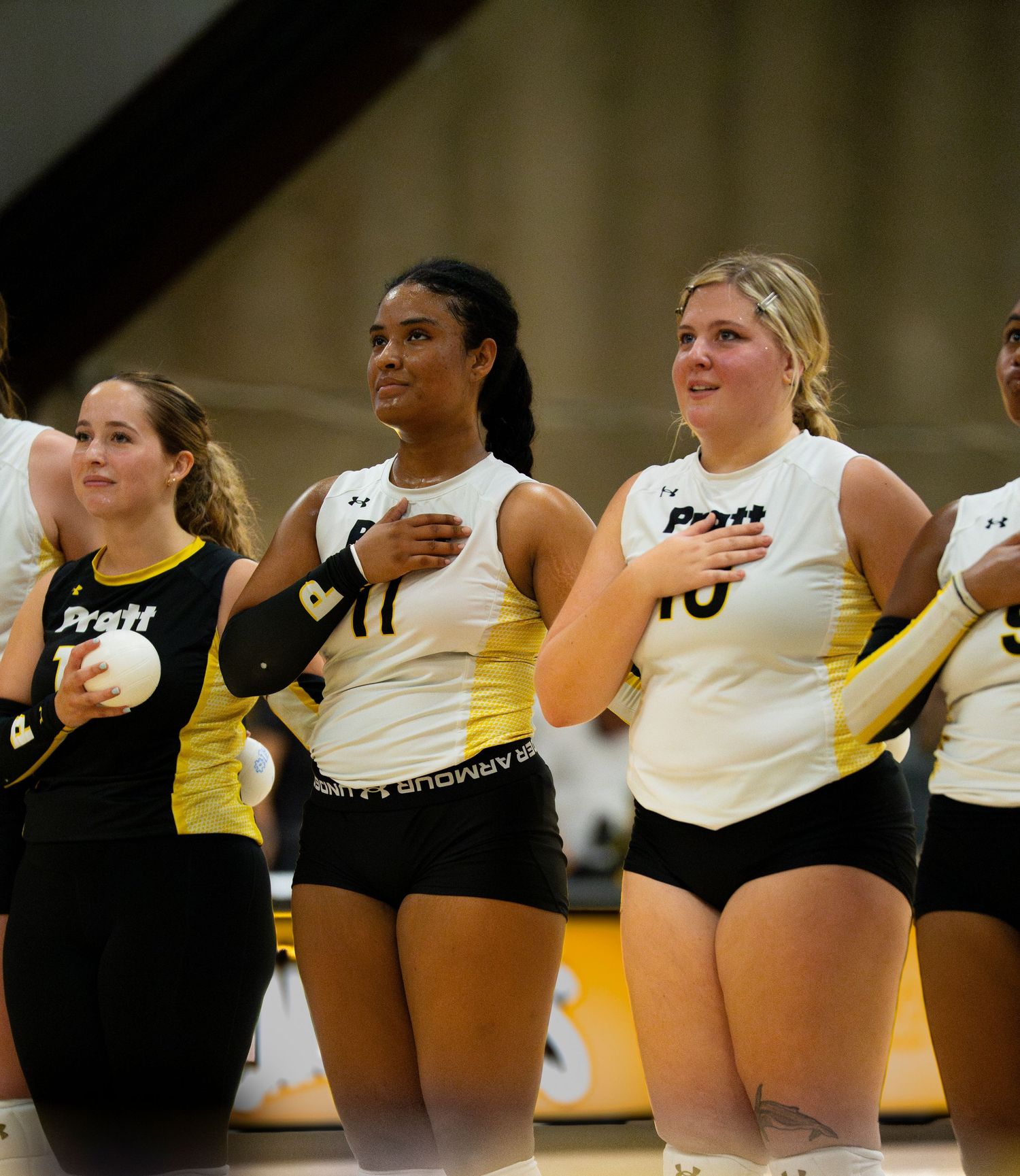 Pratt volleyball players stand in a line with hands over hearts during the national anthem before a match.