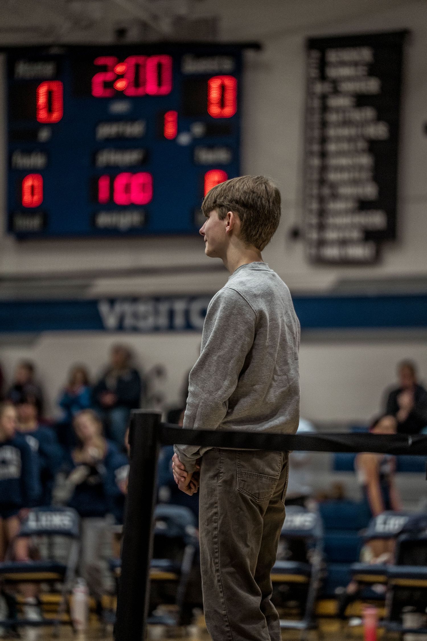 Young boy stands courtside watching a wrestling meet with a scoreboard showing 2:00 in background.