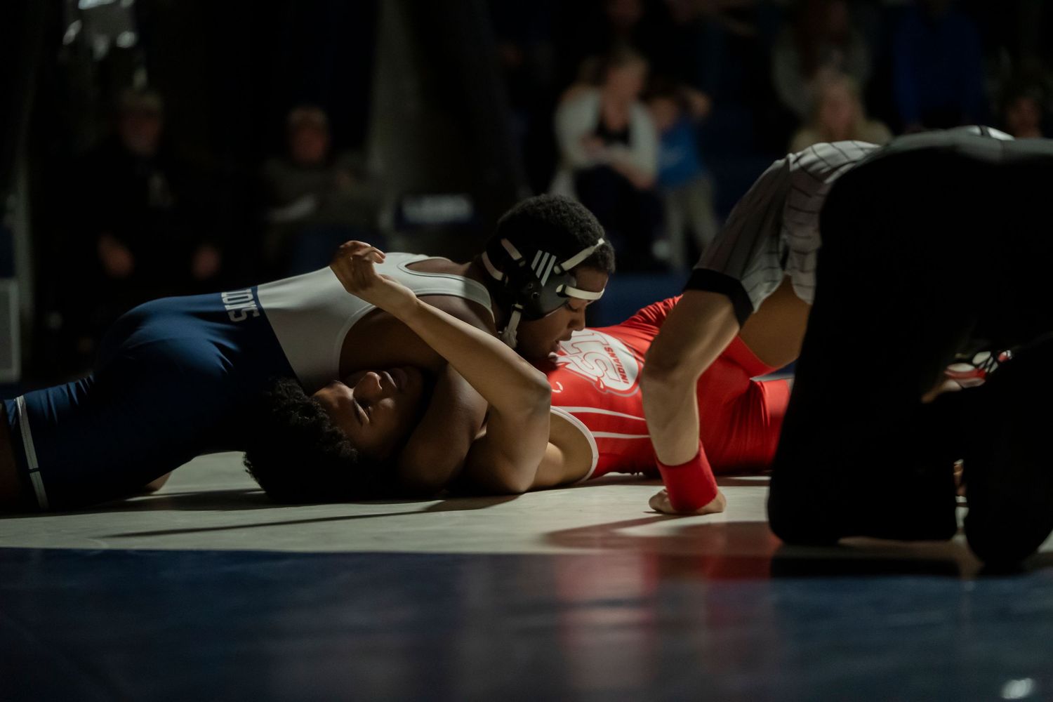 Two high school wrestlers battle on the mat as referee watches closely during an intense wrestling competition.