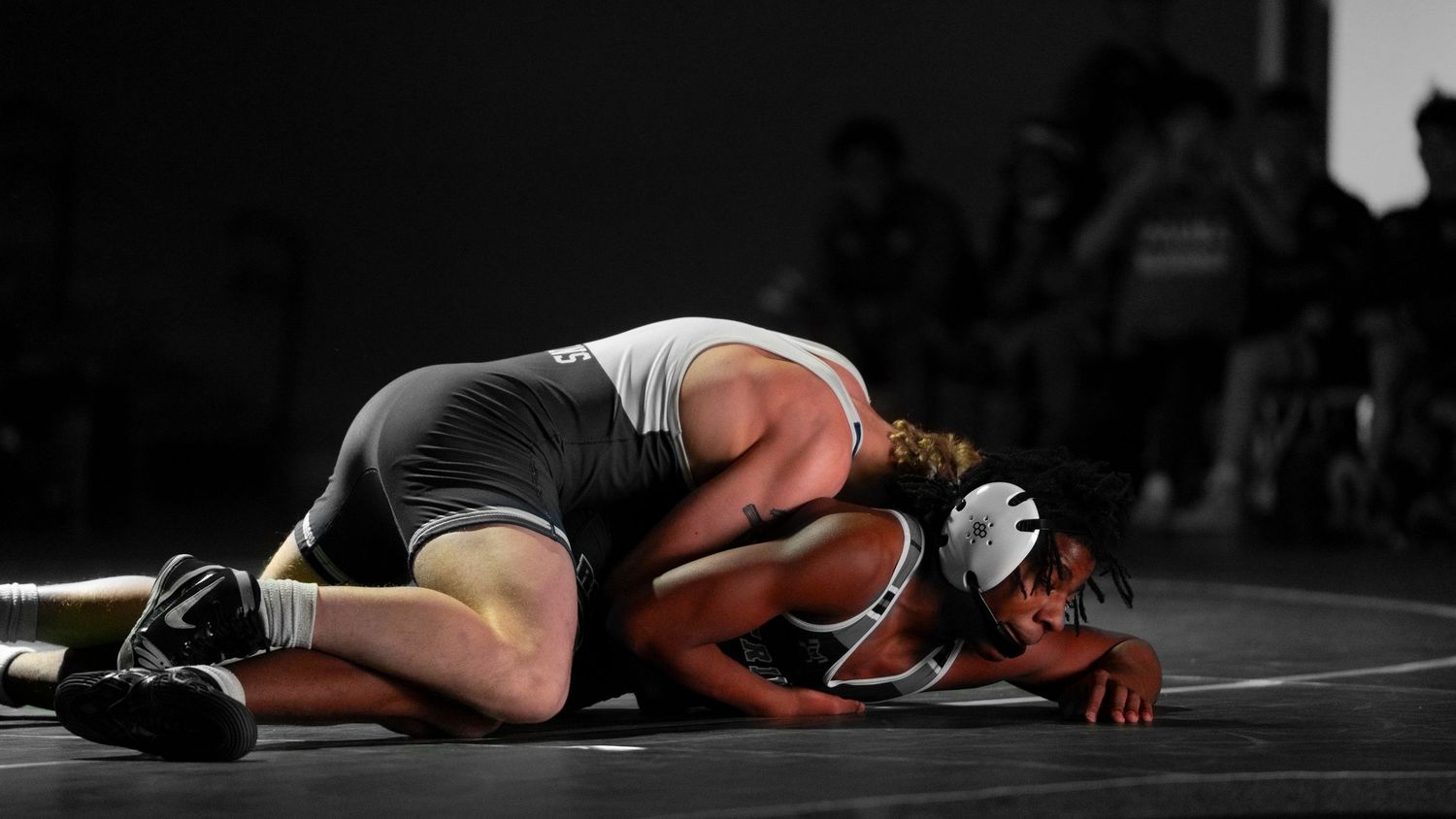 Wrestler in white singlet pins dark-uniformed opponent to the mat in a competitive wrestling match.