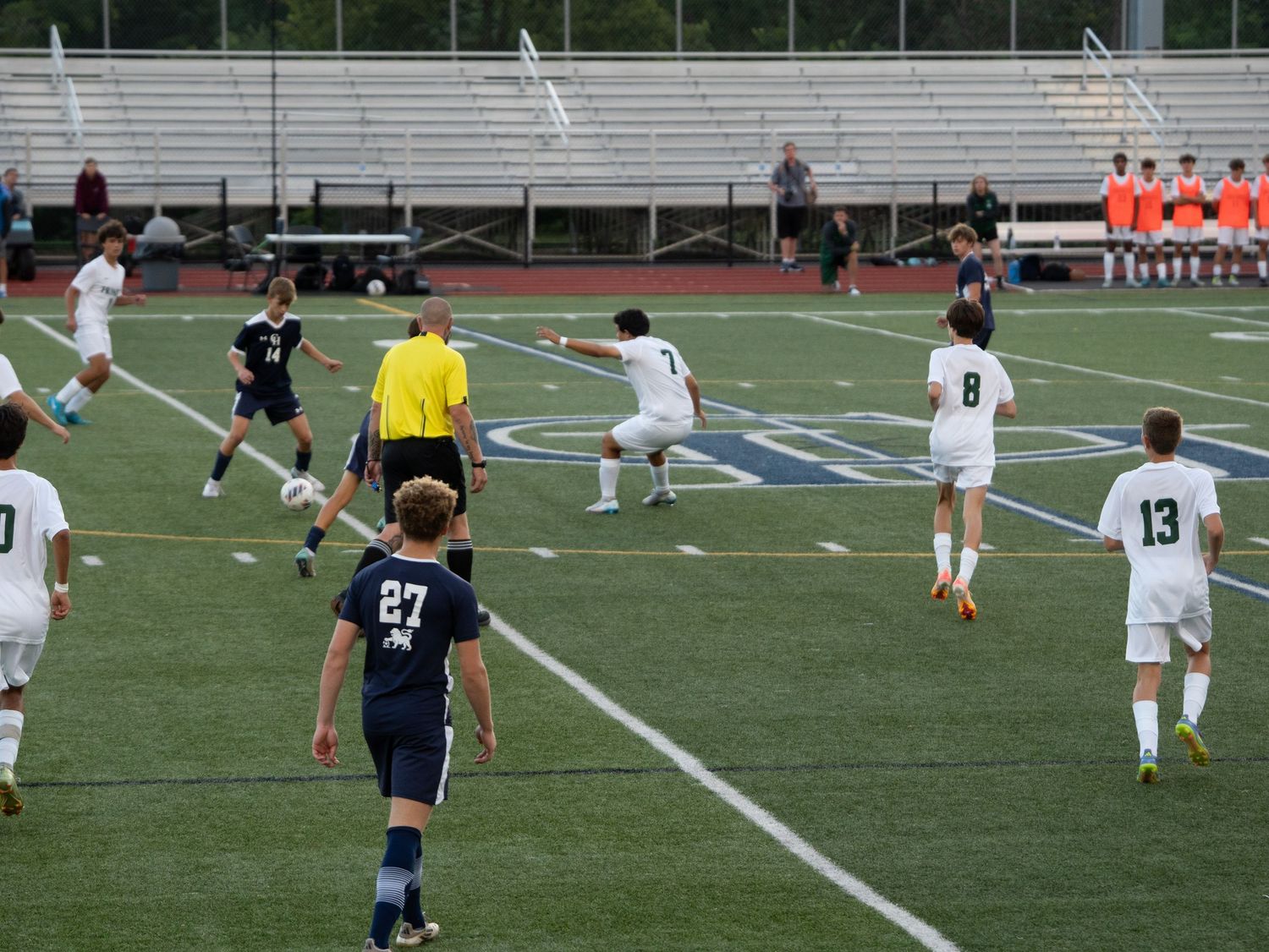 High school soccer players compete on turf field with referee in yellow during active play.