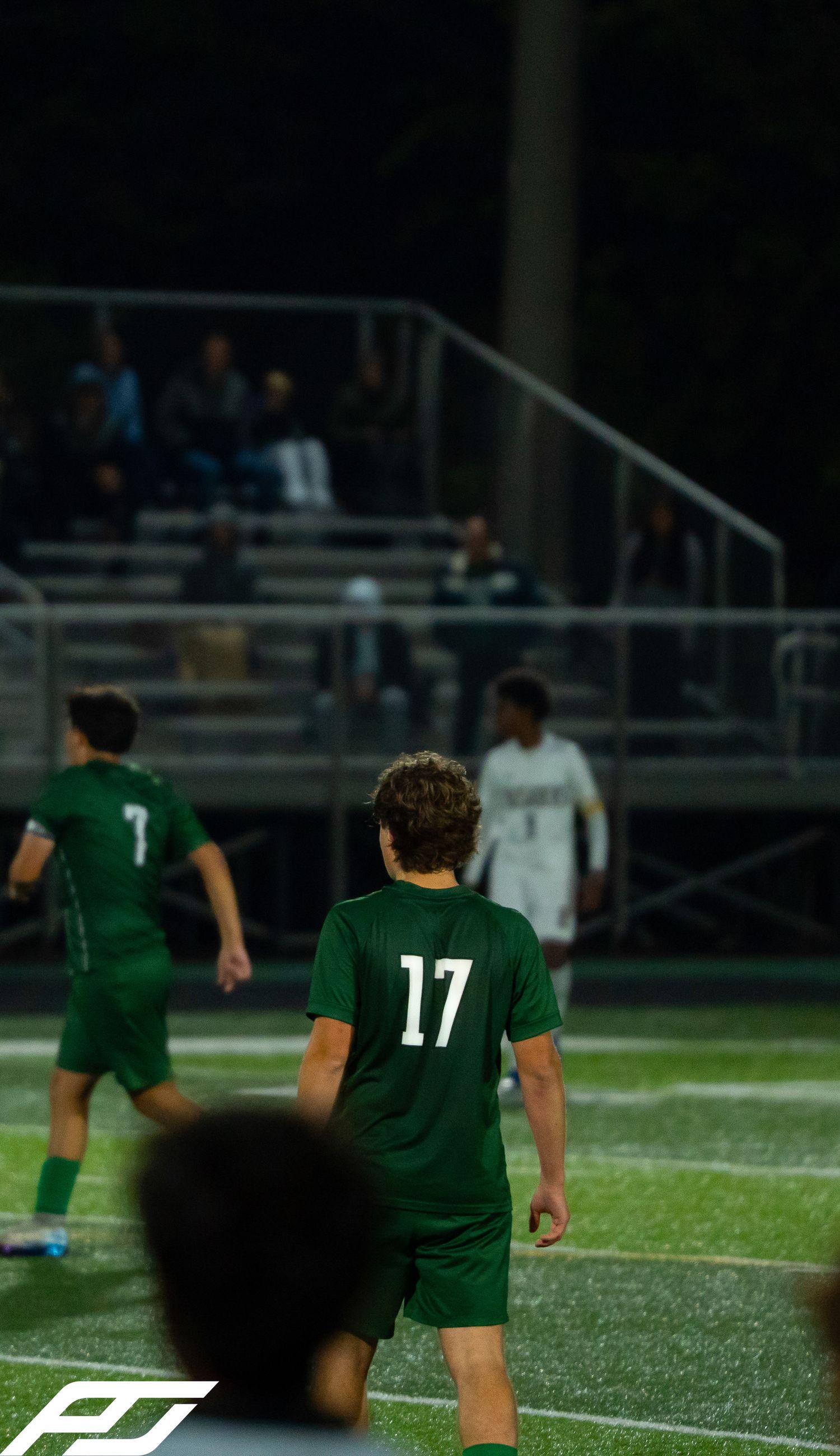 Youth soccer player wearing green number 17 jersey stands on illuminated field during a night game.