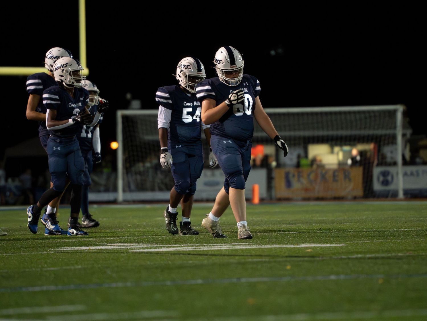 Camp Hill Lions football players in navy uniforms walking on field during a night game.