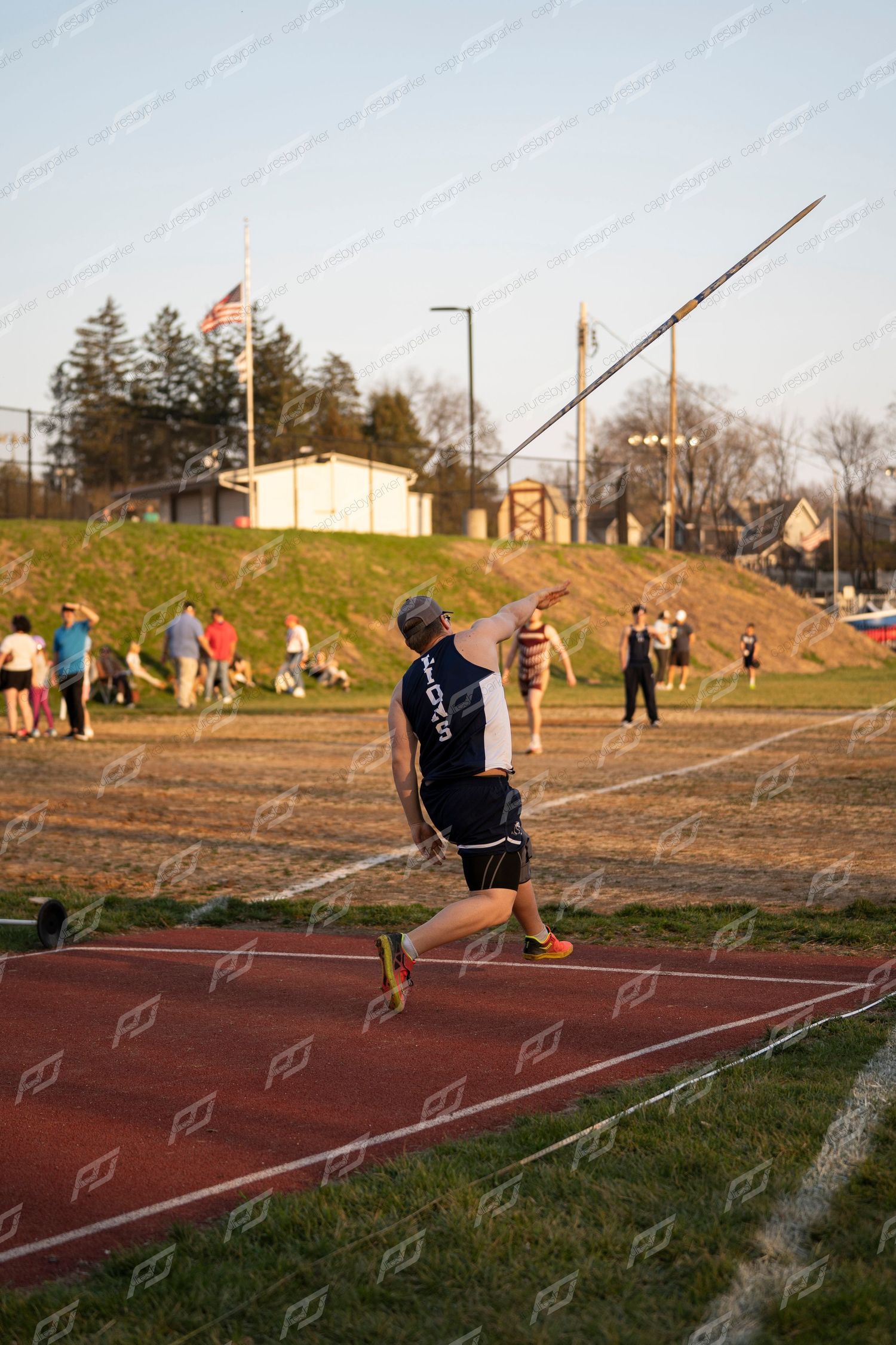 Athlete throws javelin on red track during outdoor track and field meet with American flag in background.