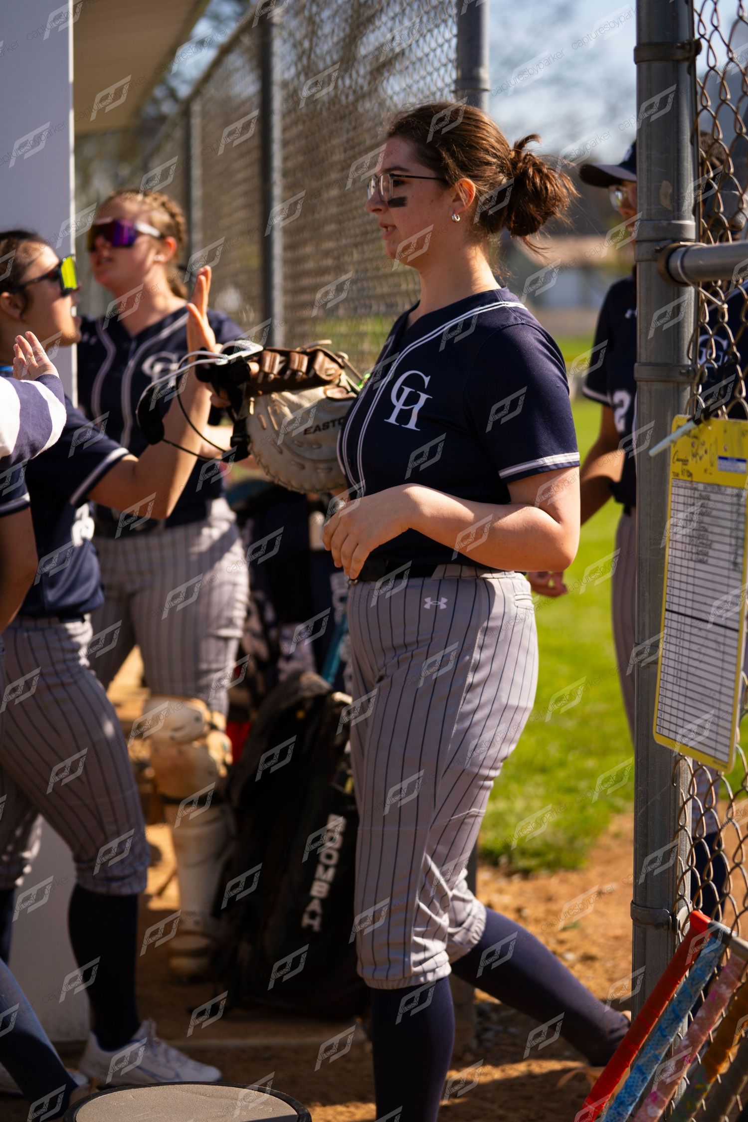 Female softball players in navy CH uniforms stand near dugout fence during game.