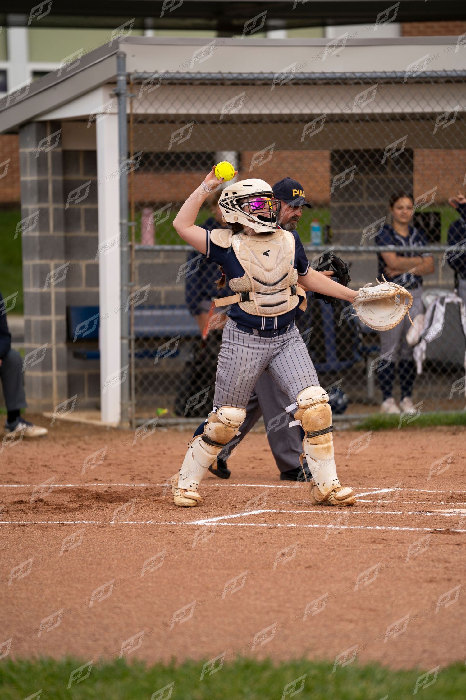 Softball catcher in navy uniform preparing to throw a yellow ball during a game.