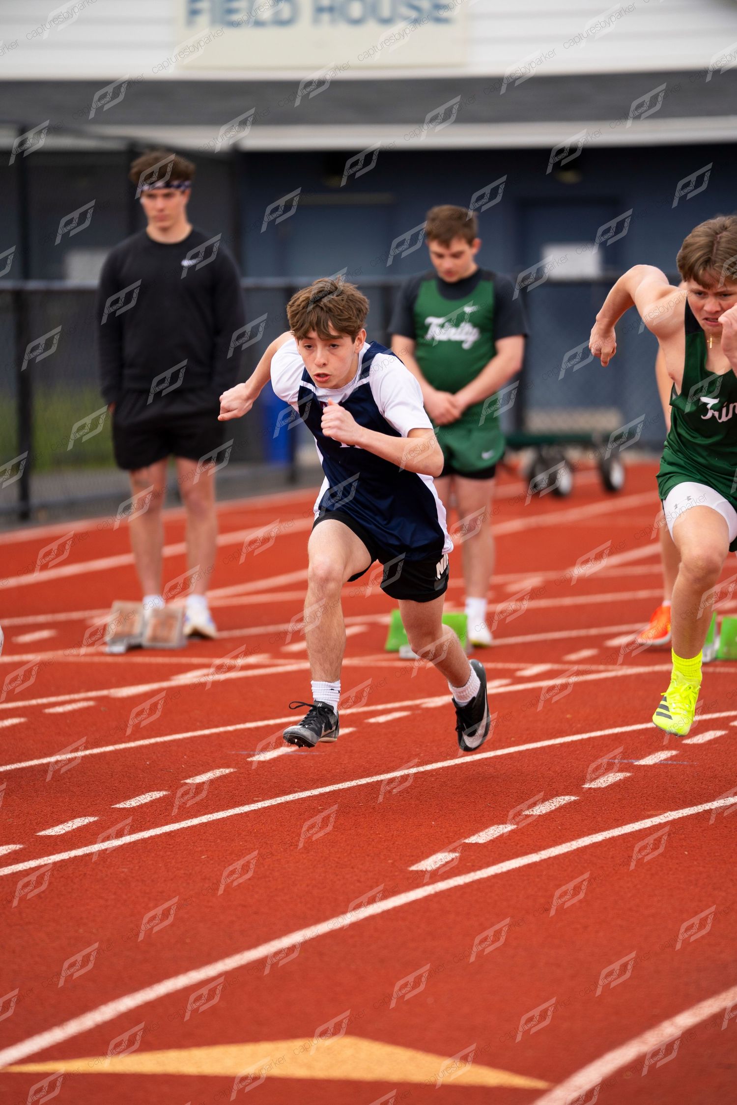 Young male sprinter in blue vest explodes off starting line on red track during high school meet.