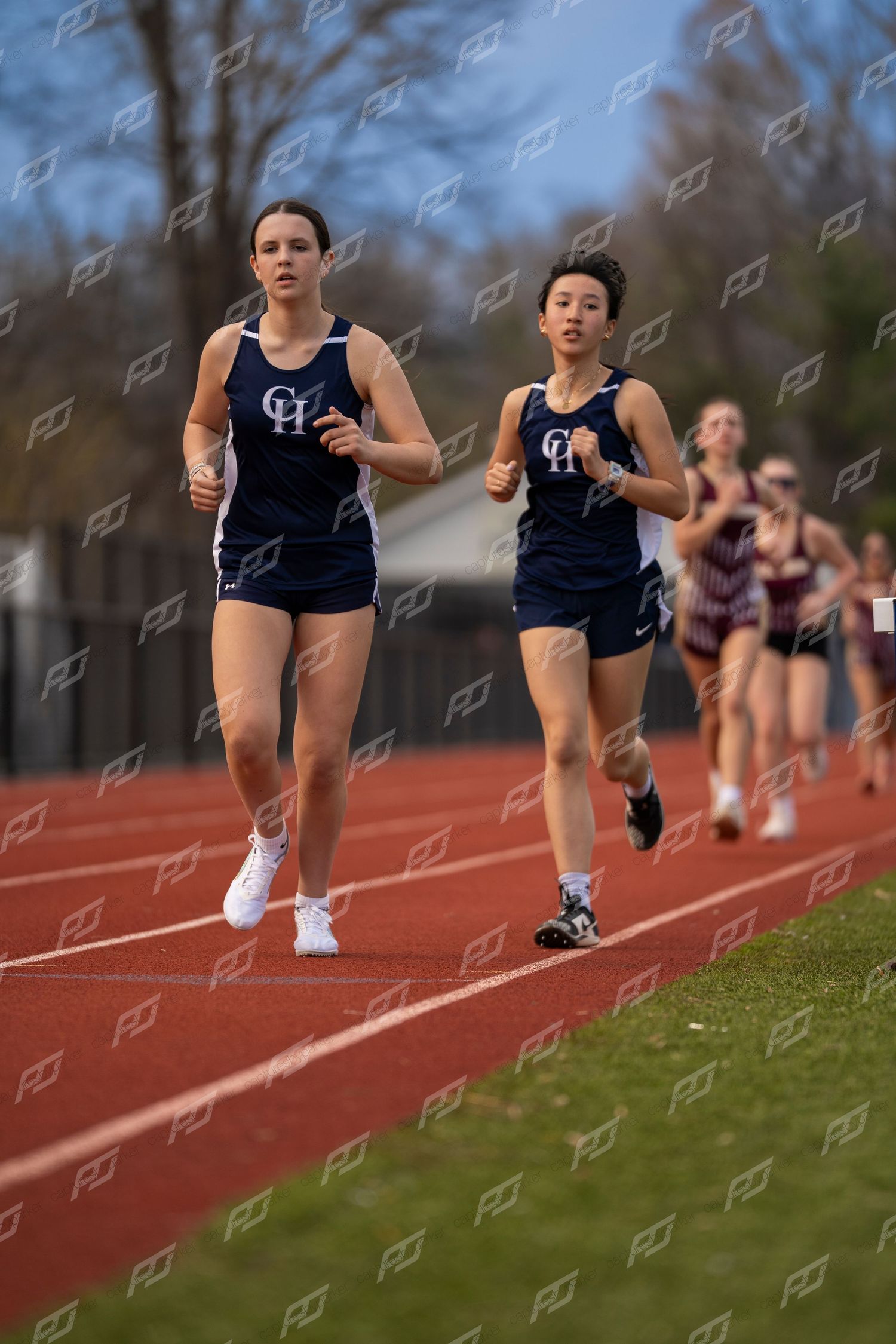 Two female runners in navy CH uniforms competing in track race on red track.