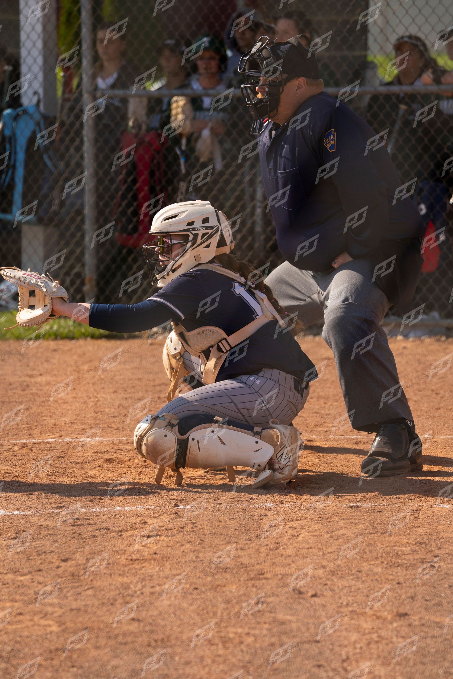 Softball catcher in full gear crouches at home plate ready to receive a pitch with umpire standing behind.