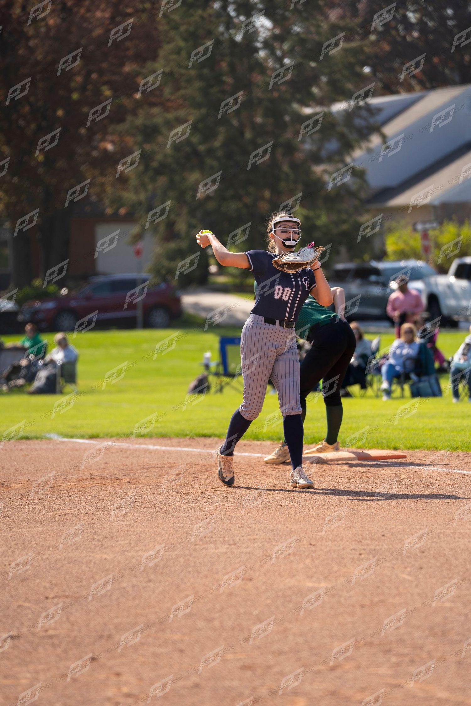 Female softball player #00 in navy pinstripe uniform throws ball from third base position.