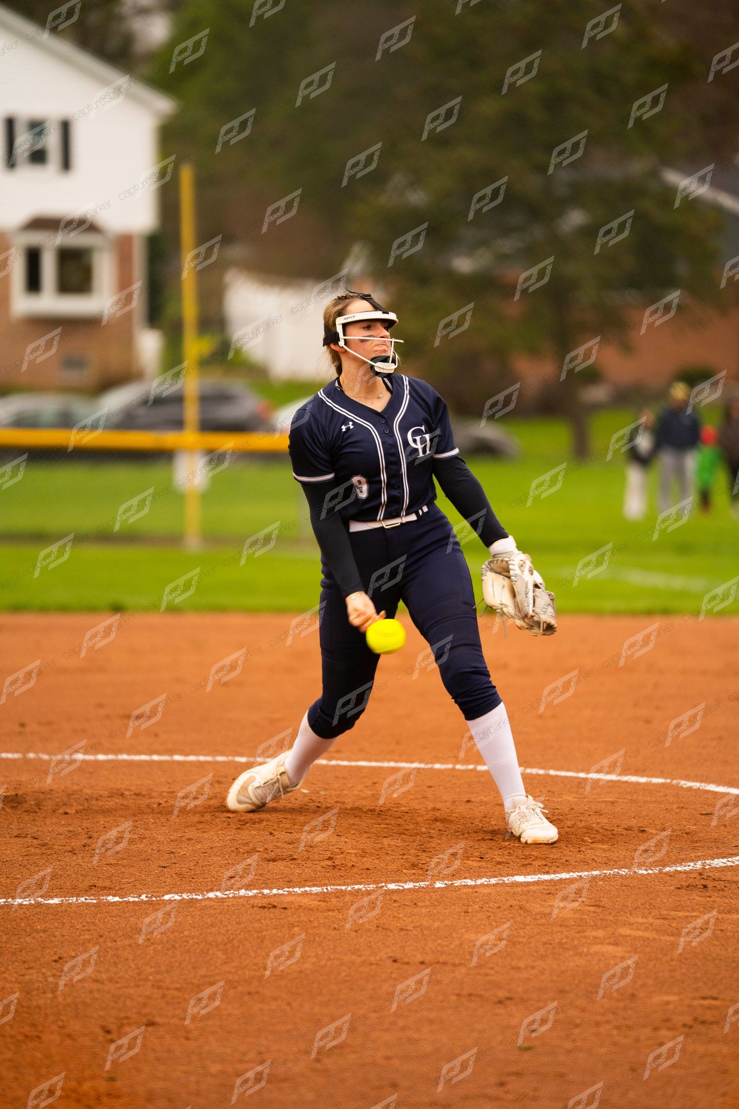 Female softball pitcher in navy uniform prepares to release a yellow ball during an outdoor game.