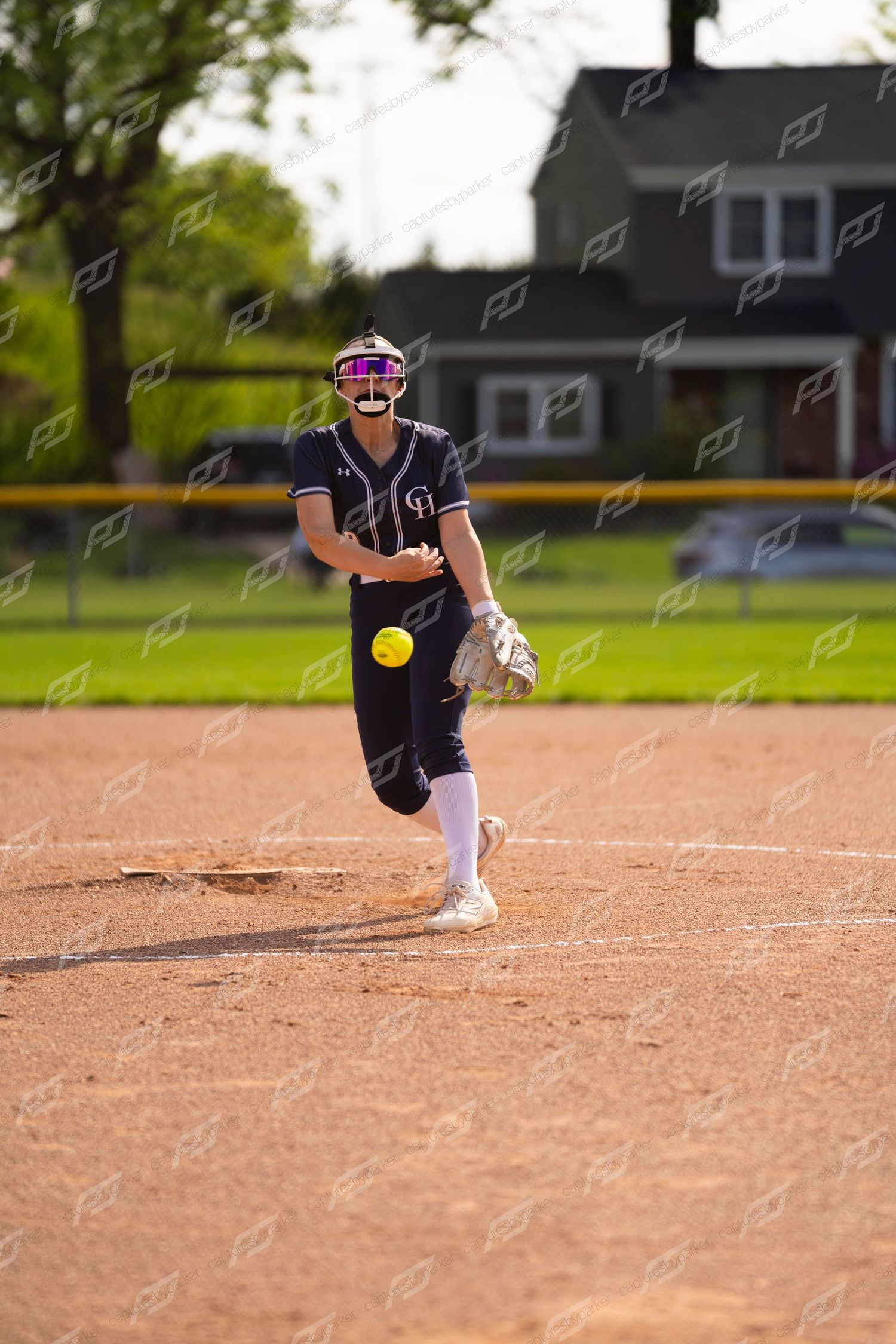 Female softball pitcher in navy uniform releases yellow ball during windmill pitch on mound.