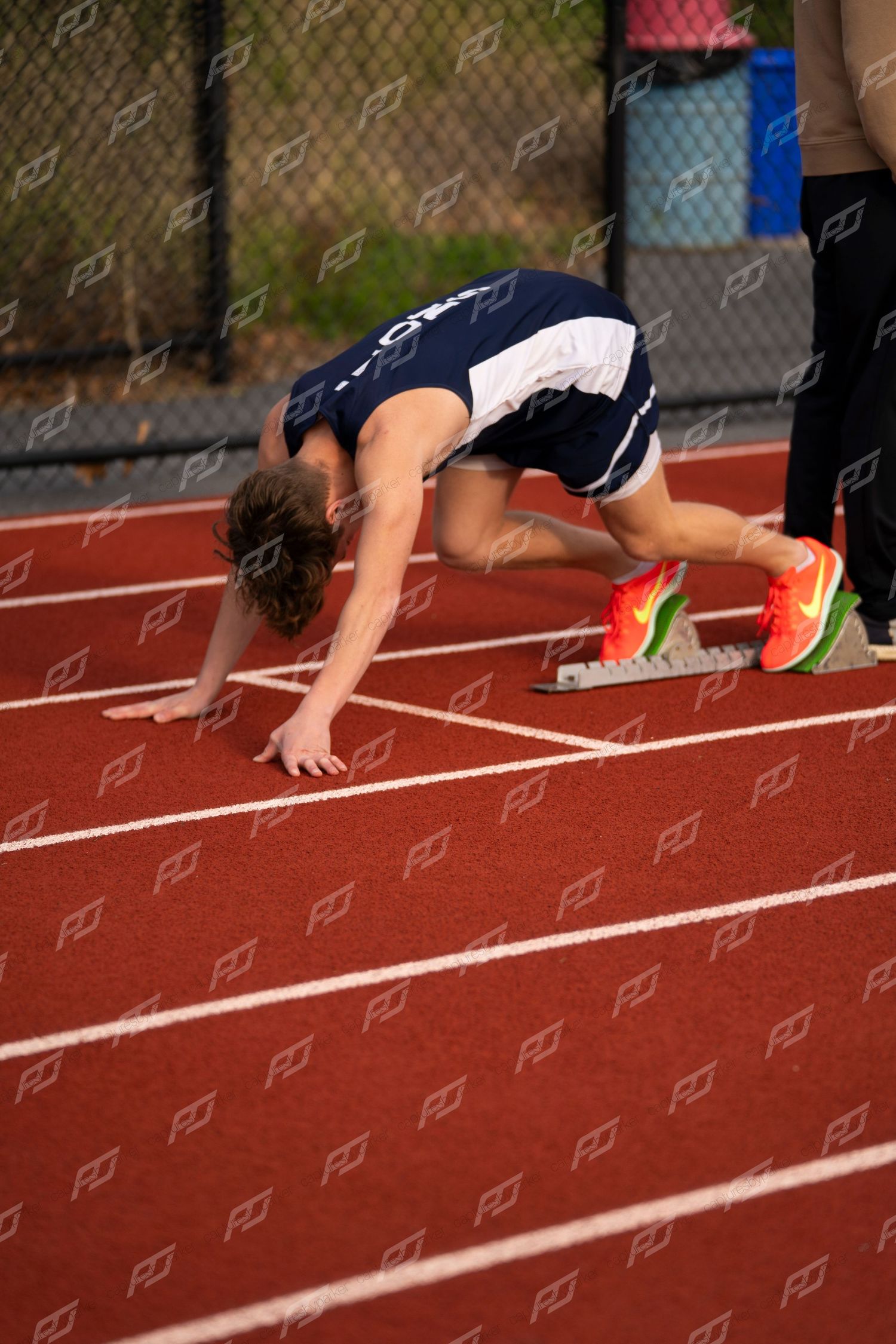 Male runner in navy uniform sets up in starting position on red track with orange starting blocks.