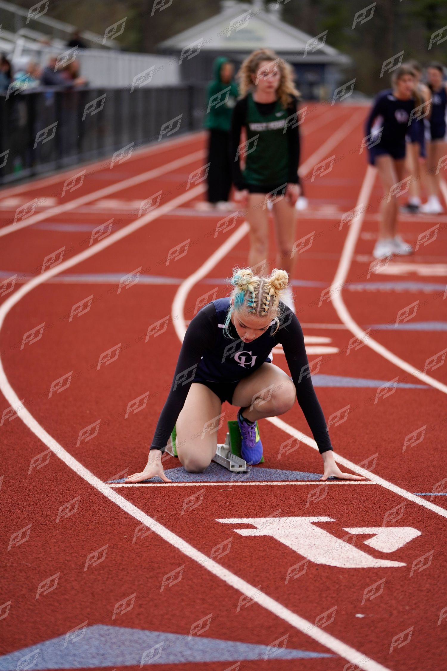 Female sprinter in starting position on a red track wearing a black uniform with colorful braided hair.