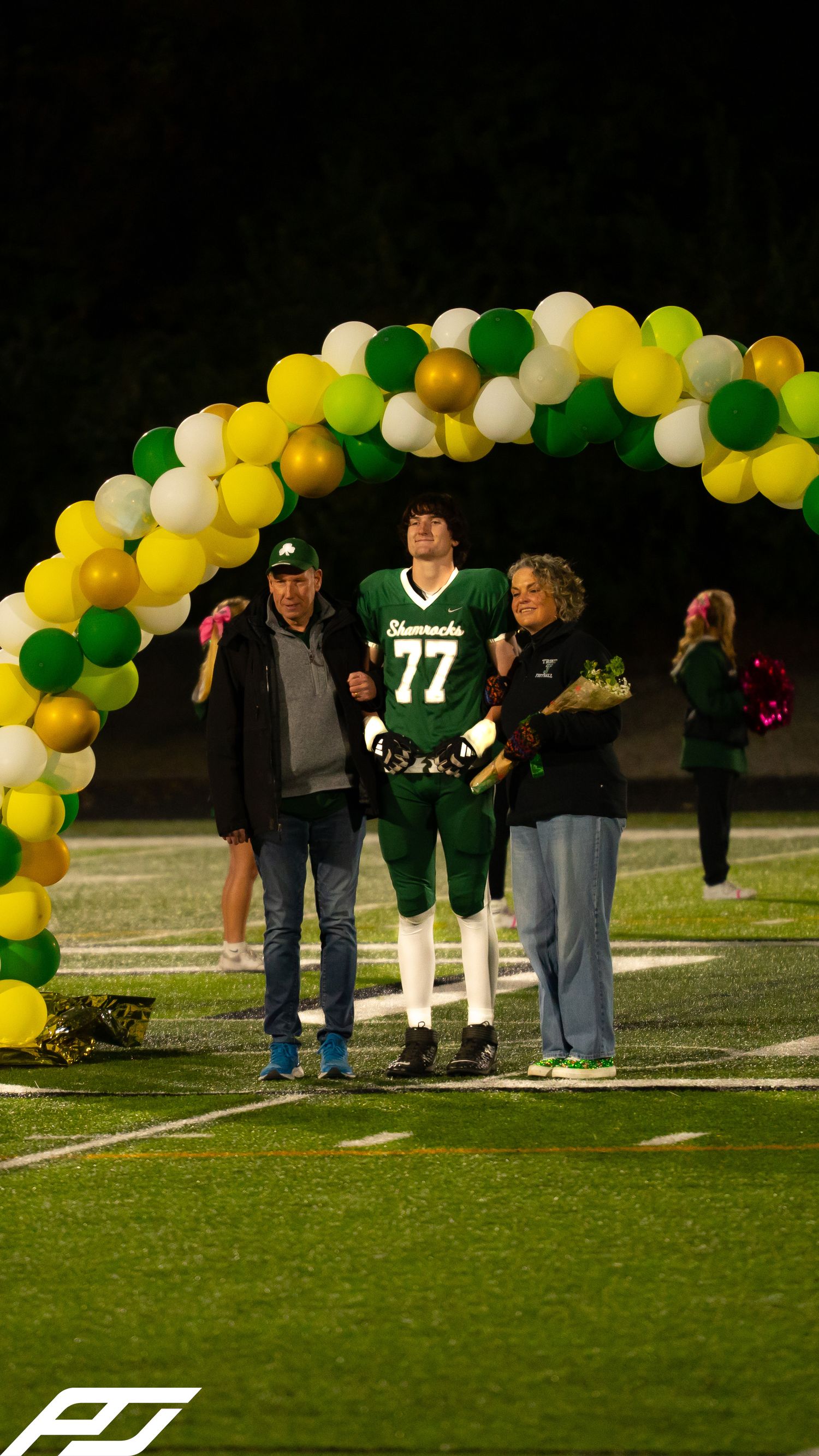 Shamrocks football player #77 stands with parents under green and gold balloon arch during senior night.