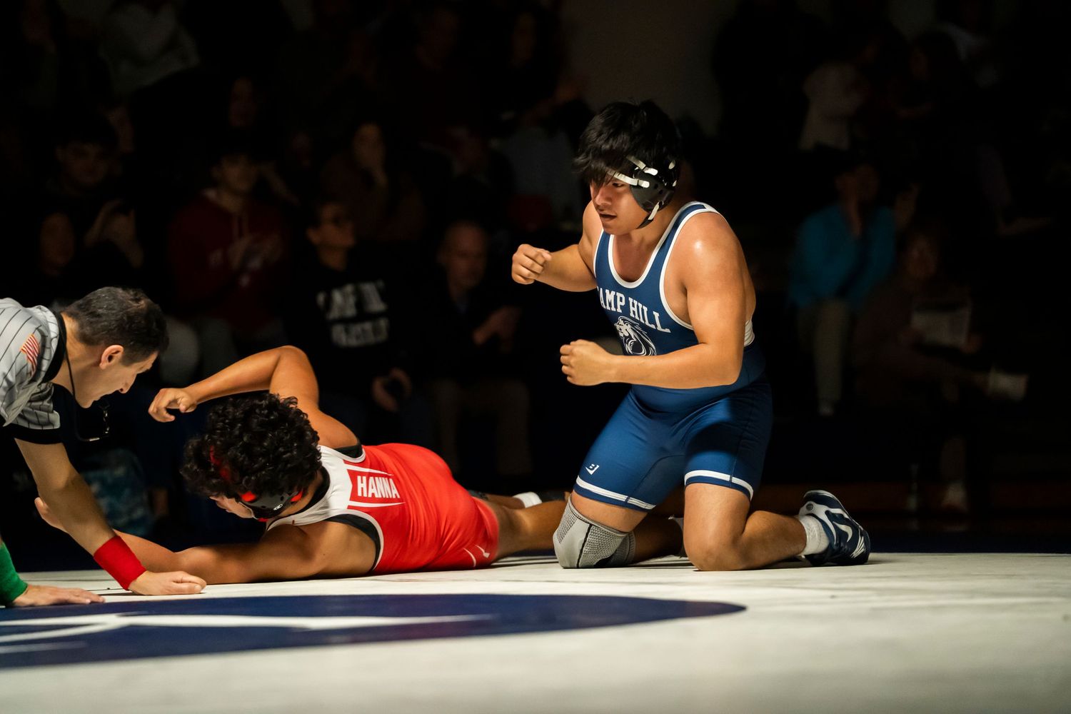 Blue singlet wrestler celebrates victory on knees as defeated opponent in red lies flat on wrestling mat.