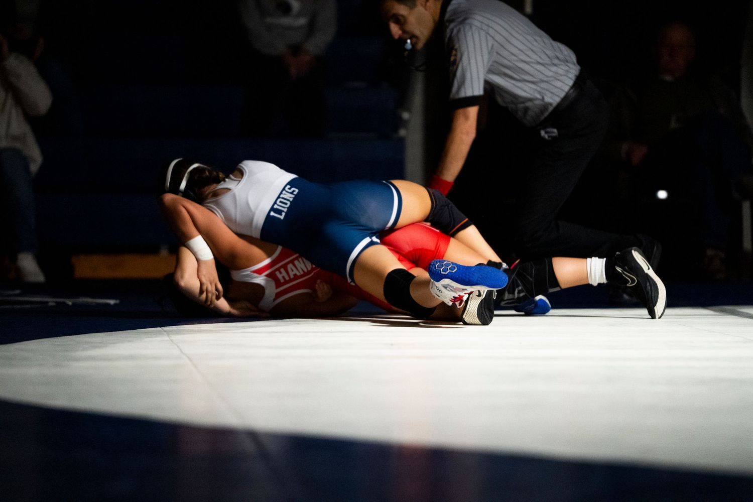 Two wrestlers compete on a blue mat during a high school wrestling match with a referee observing.