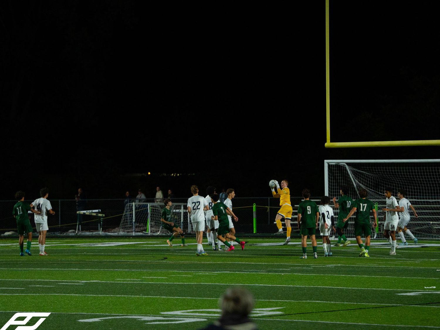 Soccer players on a turf field during a night game with a goalkeeper in yellow jersey.