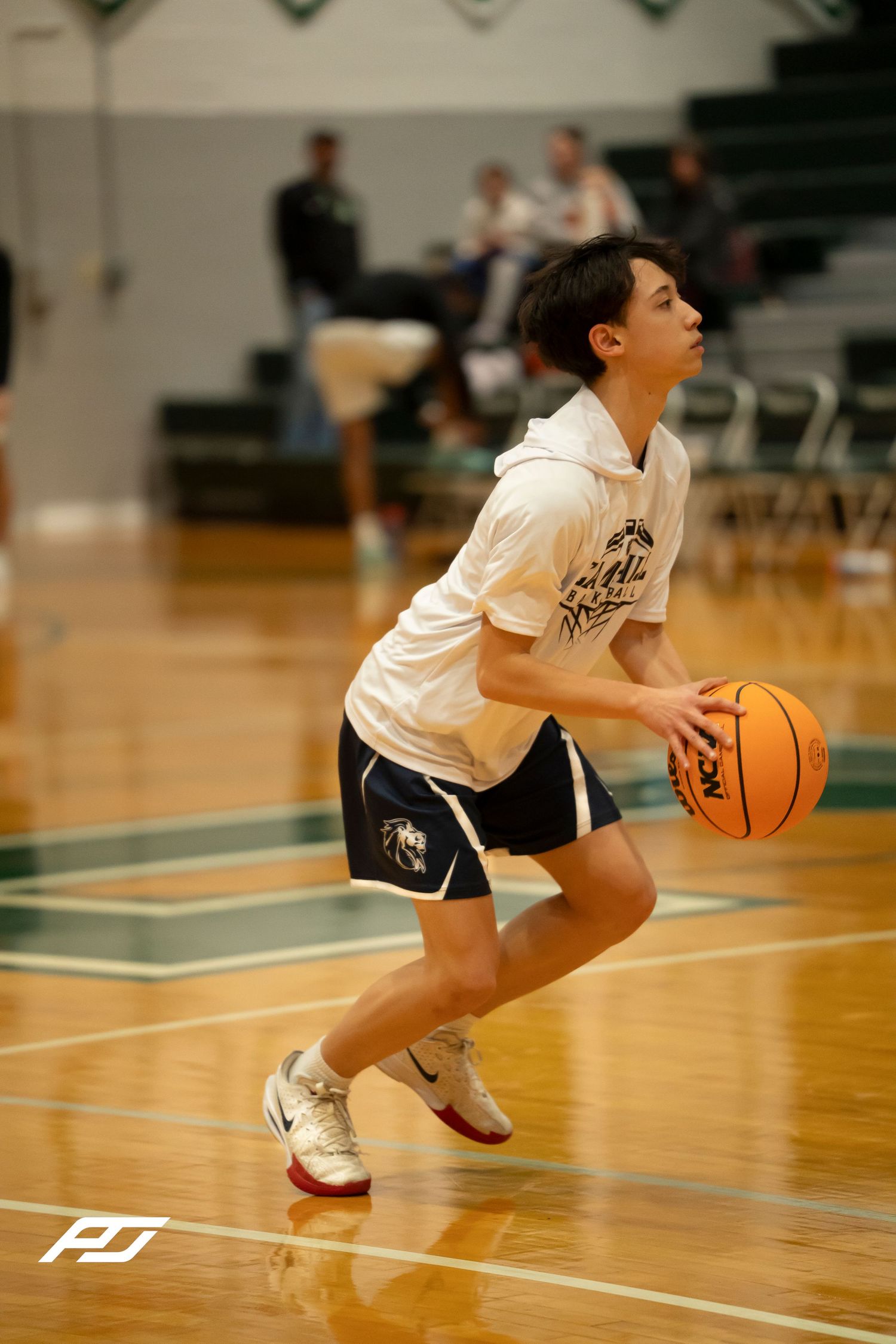 Young basketball player in white hoodie dribbles orange Wilson basketball during warmups in green gymnasium.